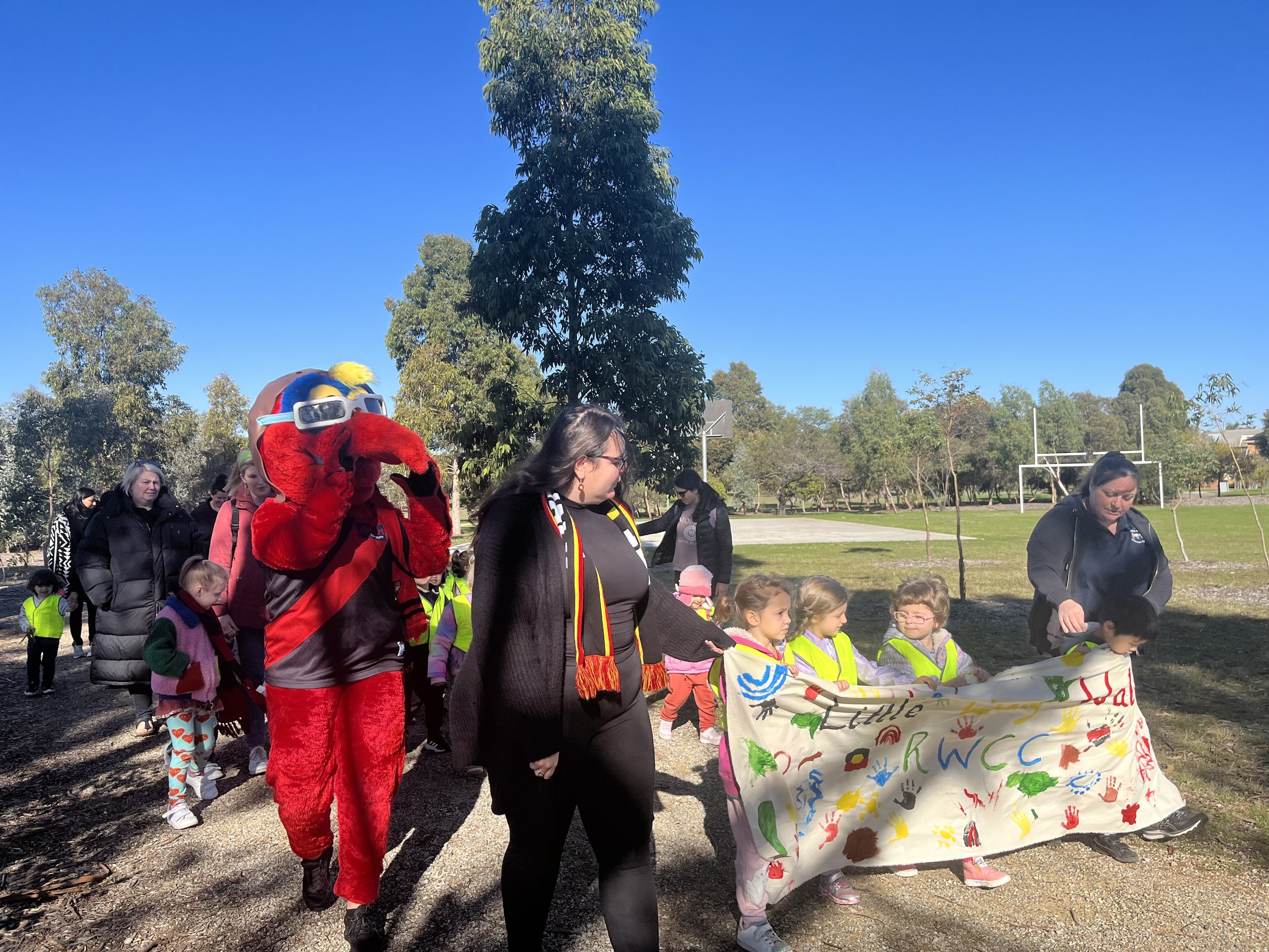 A group of children and adults participating in a community event outdoors on a sunny day. Some children are wearing bright yellow safety vests. One adult is dressed in a red mascot costume with sunglasses and yellow hair. Others are holding a colorf