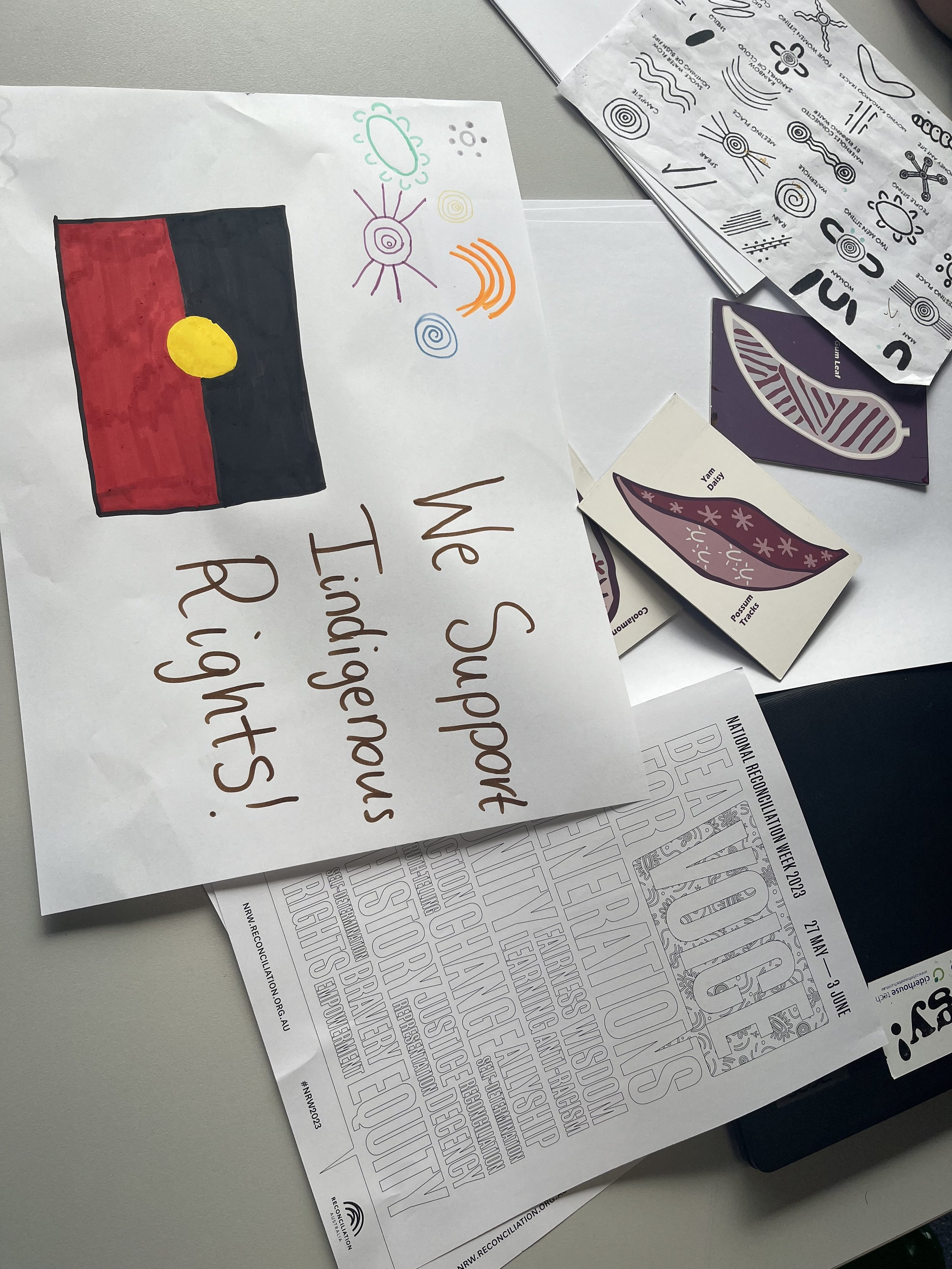 Photo of various sheets of paper on a table, including one with colored artwork of a flag, some bubble and swirl doodles, and printed worksheets with text and designs related to reconciliation week.