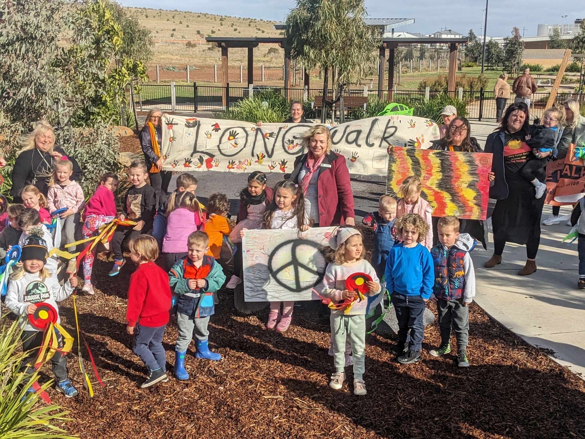 A group of children and adults gathered outdoors for a walk event, holding signs with peaceful messages, including 'Let's Walk' and peace symbols, on a sunny day in a park area with trees and structures in the background.
