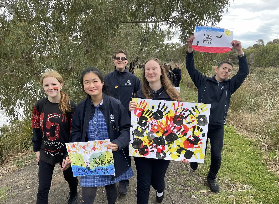 Group of five children outdoors, holding colorful paintings, with trees and a body of water in the background.