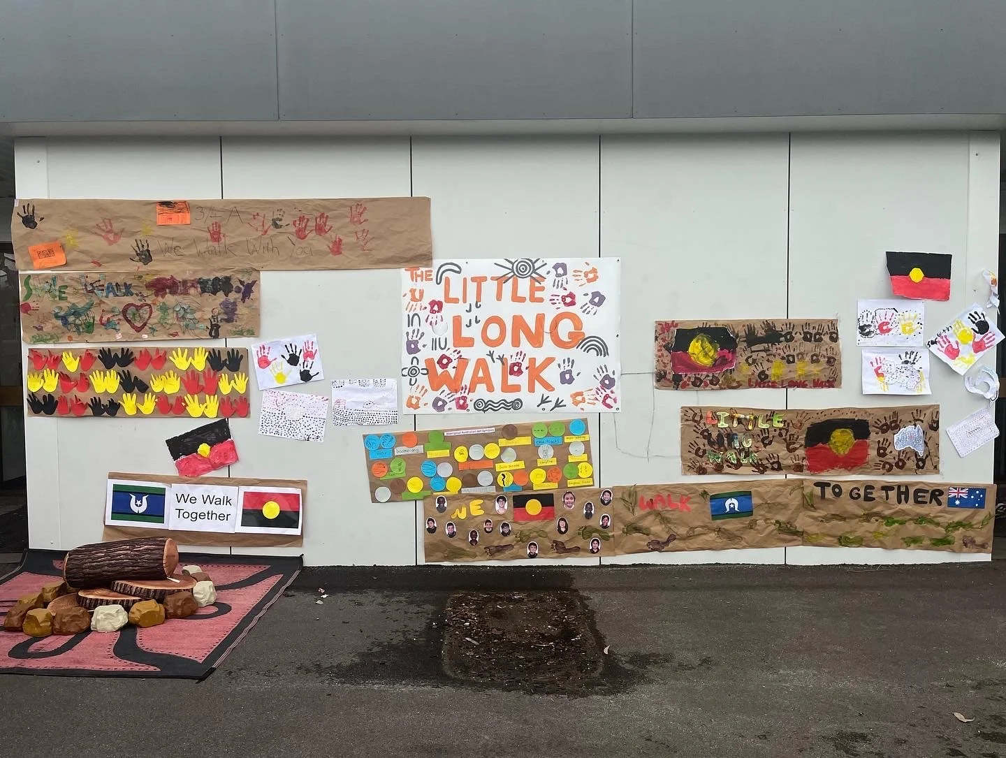 Wall decorated with artwork and banners for a community walk event, with messages such as 'The Little Long Walk' and images of handprints, flags, and painted designs. There is a log and rocks on a rug on the ground in front of the wall.