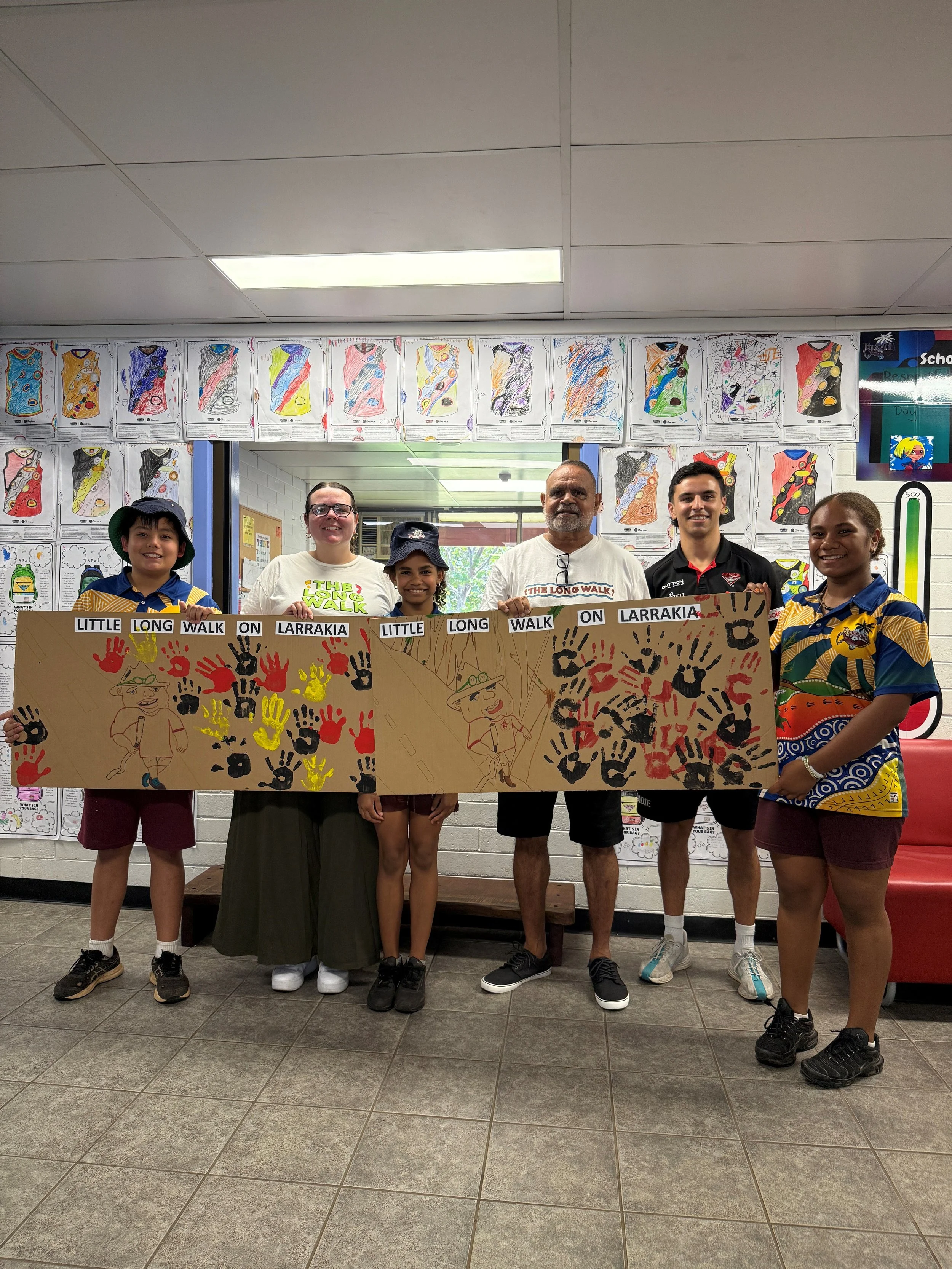 Group of six people, four children and two adults, standing indoors behind a large handmade banner with handprints and cartoon character illustrations, celebrating the Long Walk on Larrakia.