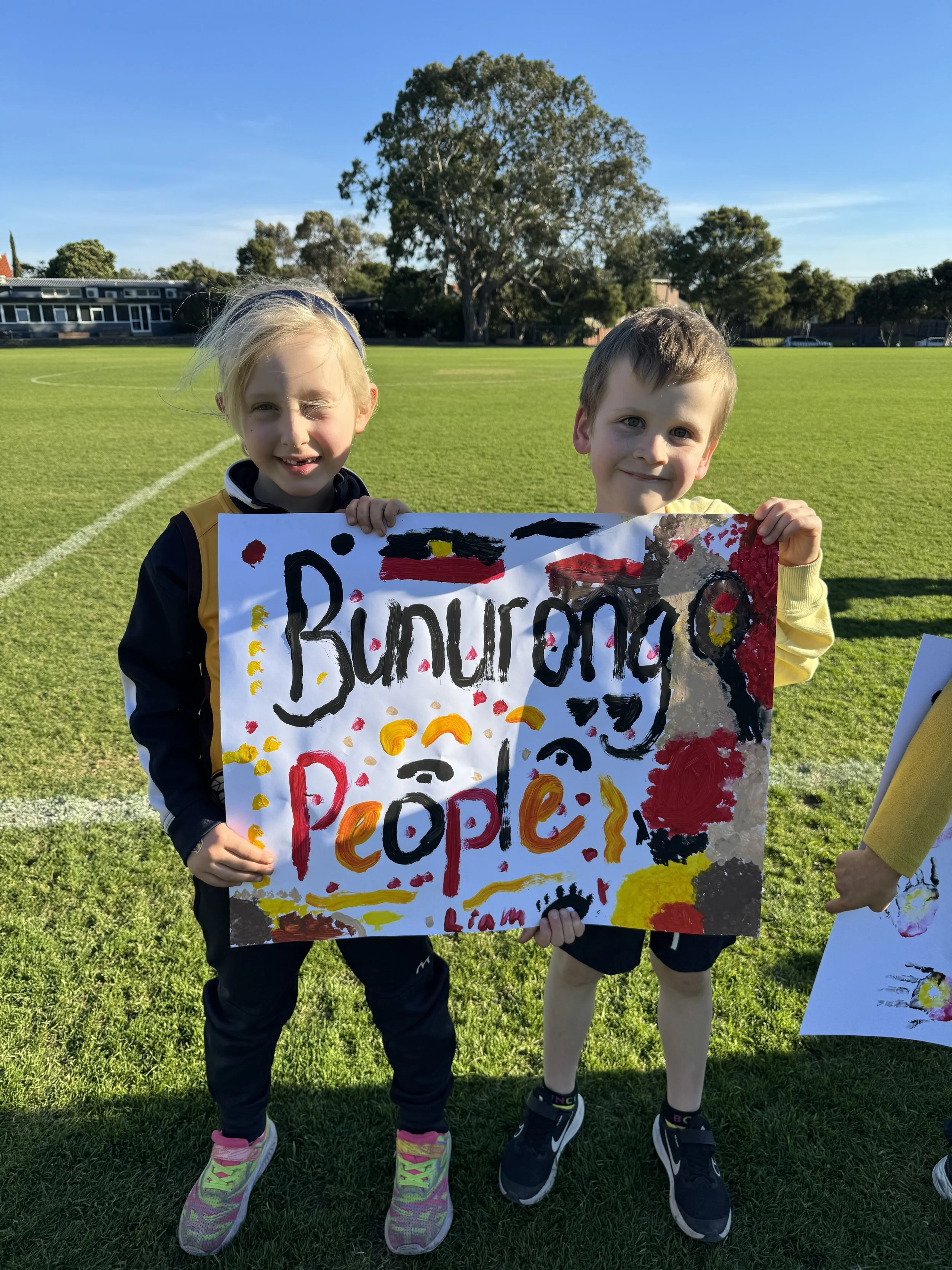 Two children standing outdoors on a grassy field, holding a colorful sign with the words 'Refugee Pops' written on it, with trees and a blue sky in the background.