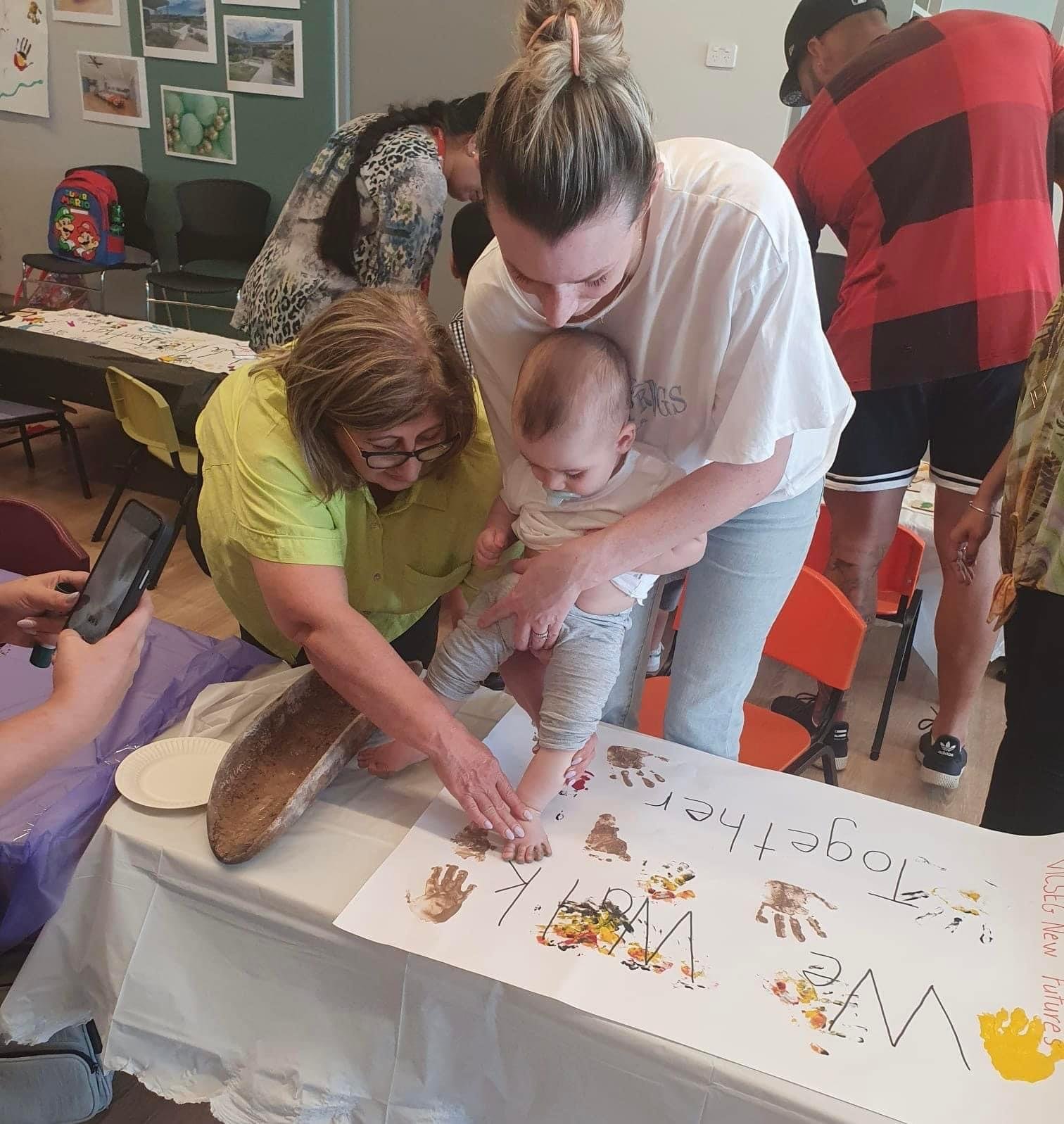A group of people, including a child, are gathered around a table making handprints and footprints on a large sheet of paper as part of a paint activity. The adult woman helps the child press their hand onto the paper, which is decorated with various