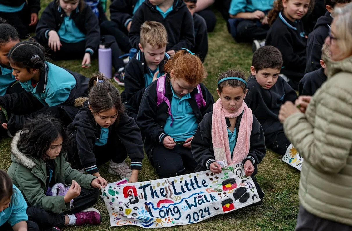 Children sitting on the grass during an outdoor event, holding a colorful handmade banner that reads 'The Little Long Walk 2024,' with an adult standing nearby.