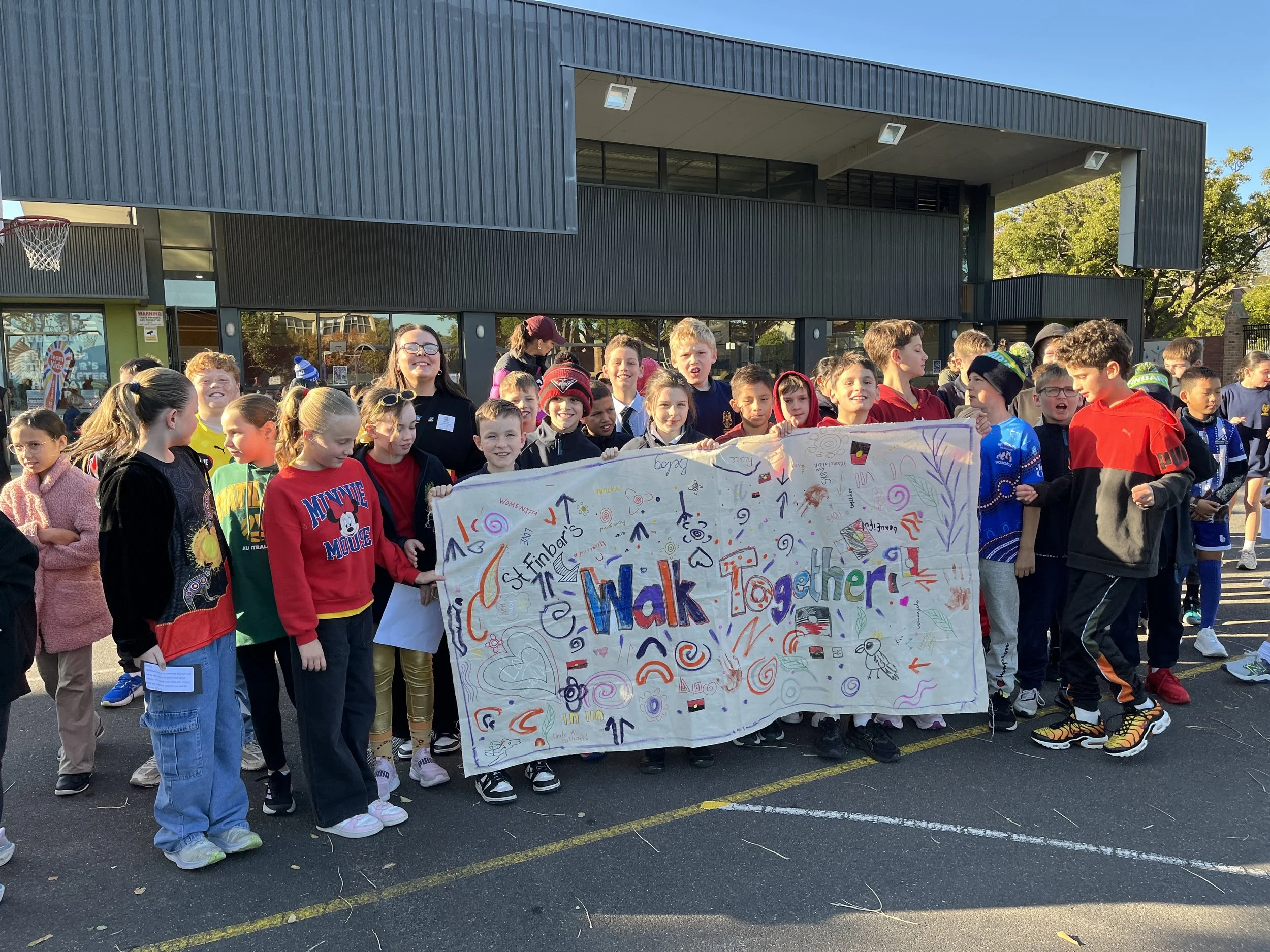 A group of children and a woman holding a large, colorful banner that reads 'Walk Together.' The children are smiling and standing outdoors in front of a modern building with trees in the background.