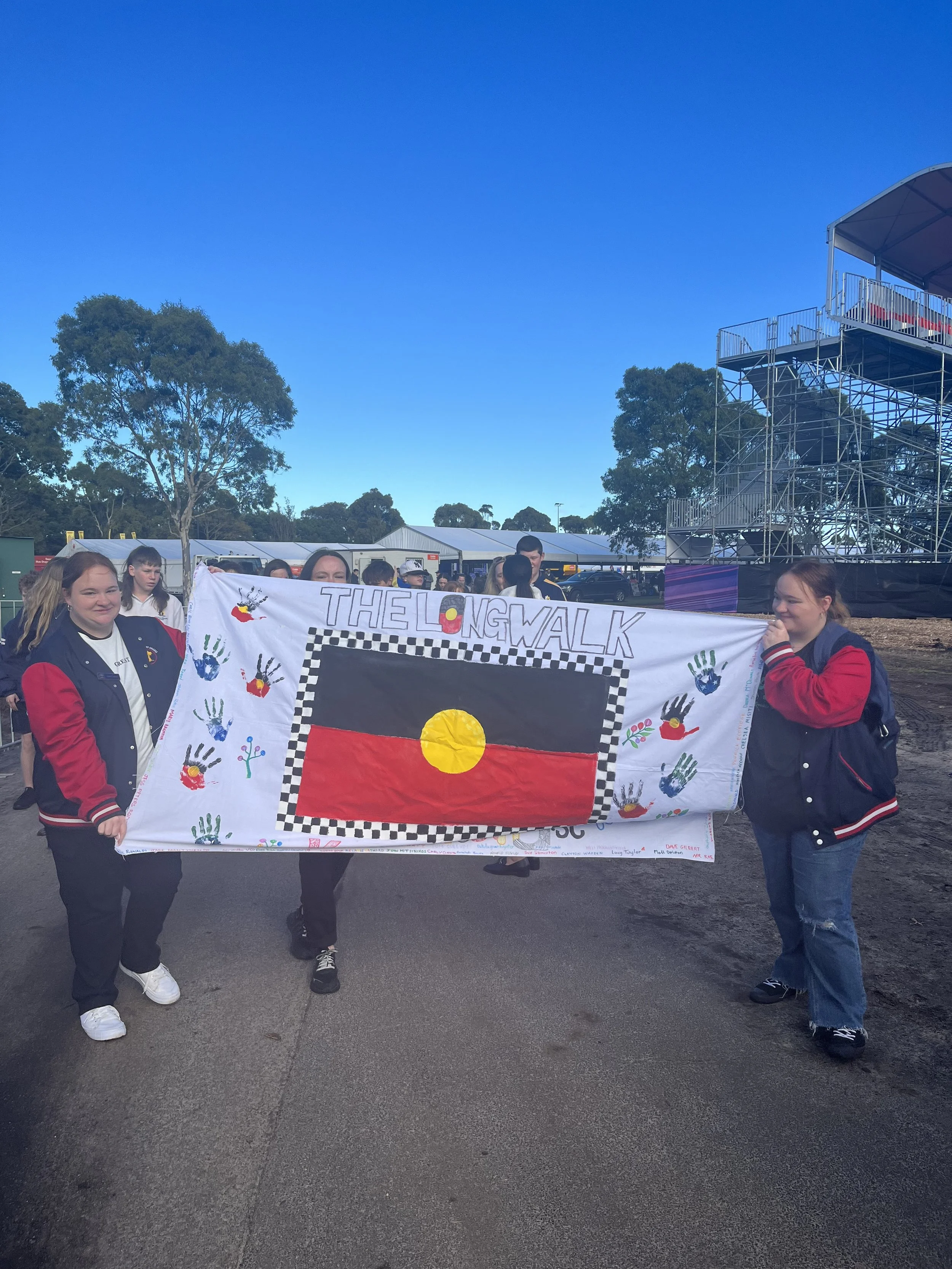 Group of young people holding a large painted banner with Australian Aboriginal art and flag motifs, outdoors under clear blue sky, with tents and trees in the background.
