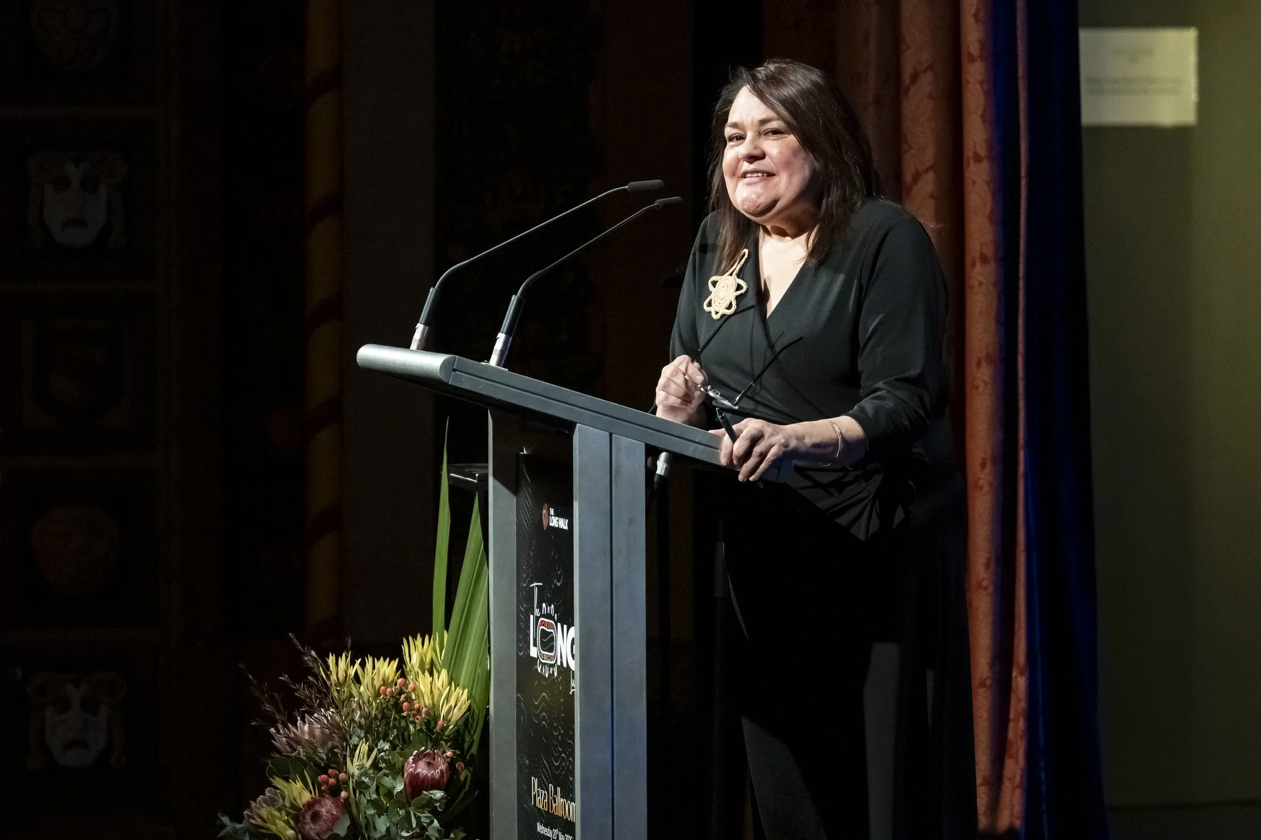 A woman standing at a podium with two microphones, holding glasses, and smiling. There is a flower arrangement in front of the podium, and a dark wooden background with a curtain on the right side.
