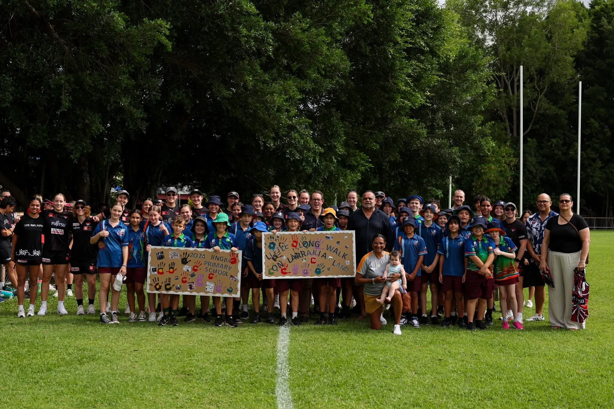 Group of children and adults posing outdoors on a sports field, holding signs with handprints and writing, surrounded by green trees.