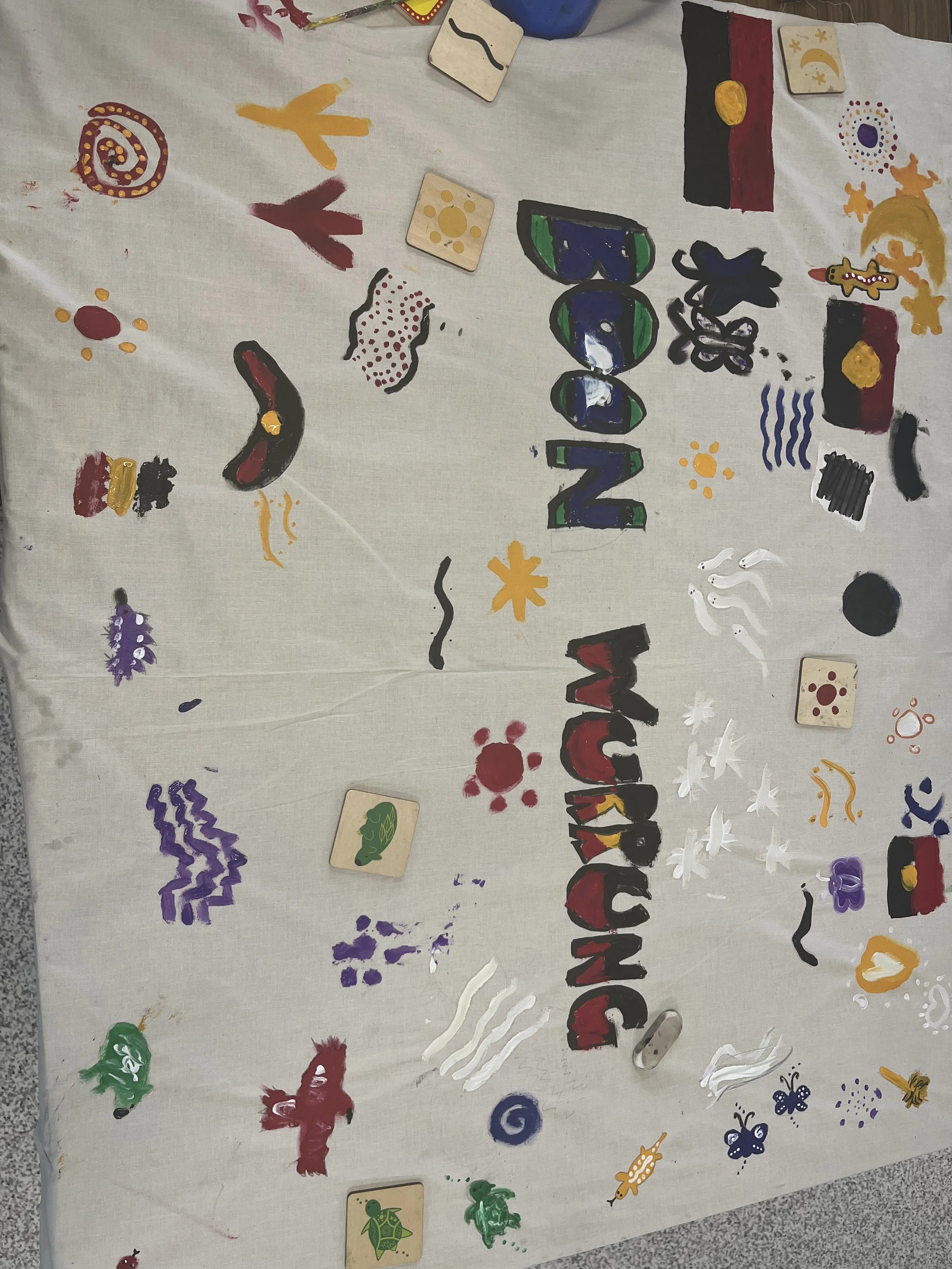 A white tablecloth with colorful hand-painted designs, including the phrase 'Welcome to the Zulu' in large colorful letters, along with various abstract shapes, animals, and symbols. There are also small tiles with animal and nature illustrations sca