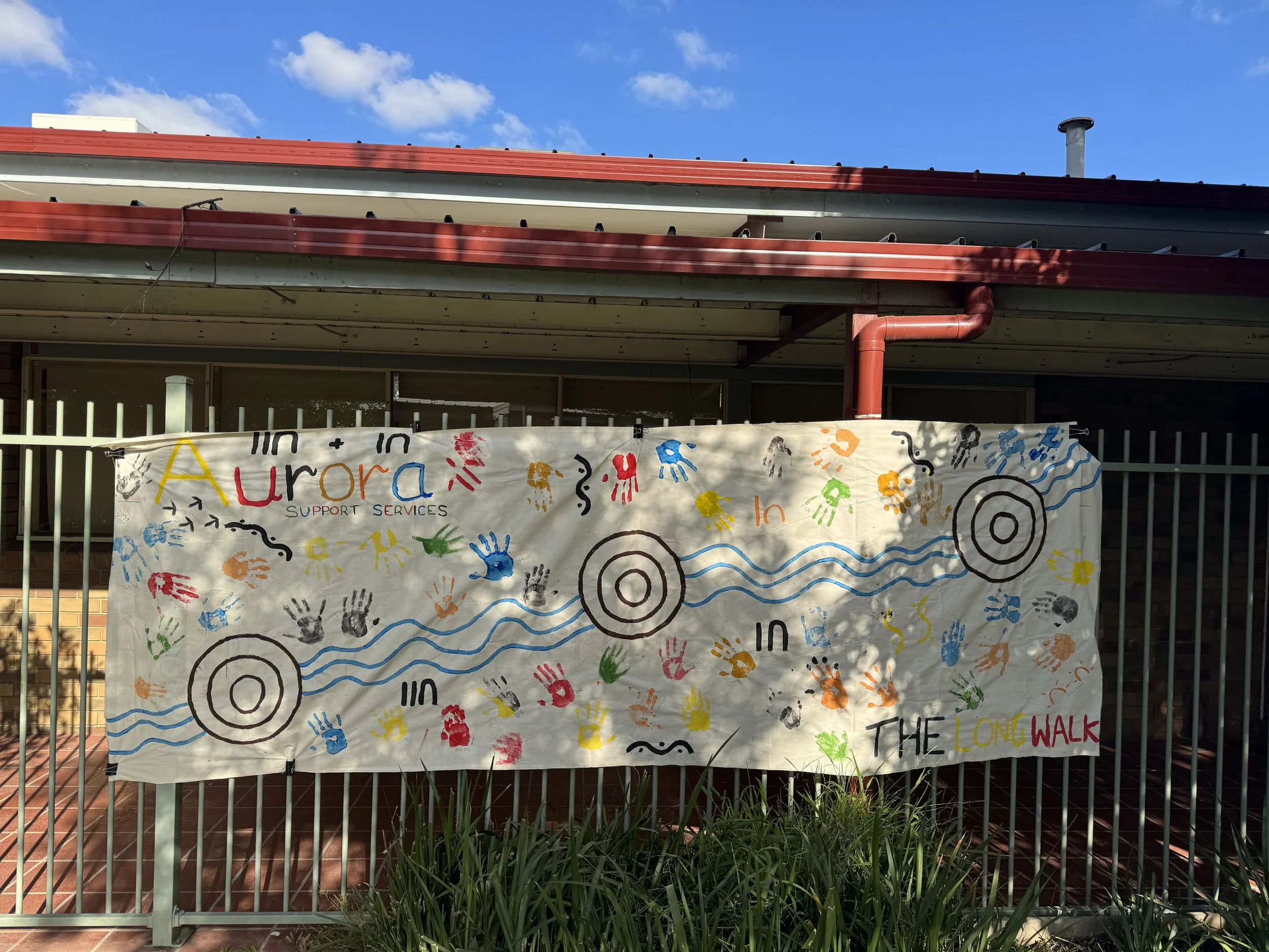 A colorful banner hangs on a fence in front of a building with a red roof and a chimney. The banner is decorated with handprints, target symbols, and the words 'Aurora support services,' 'in the long walk,' and other colorful text.