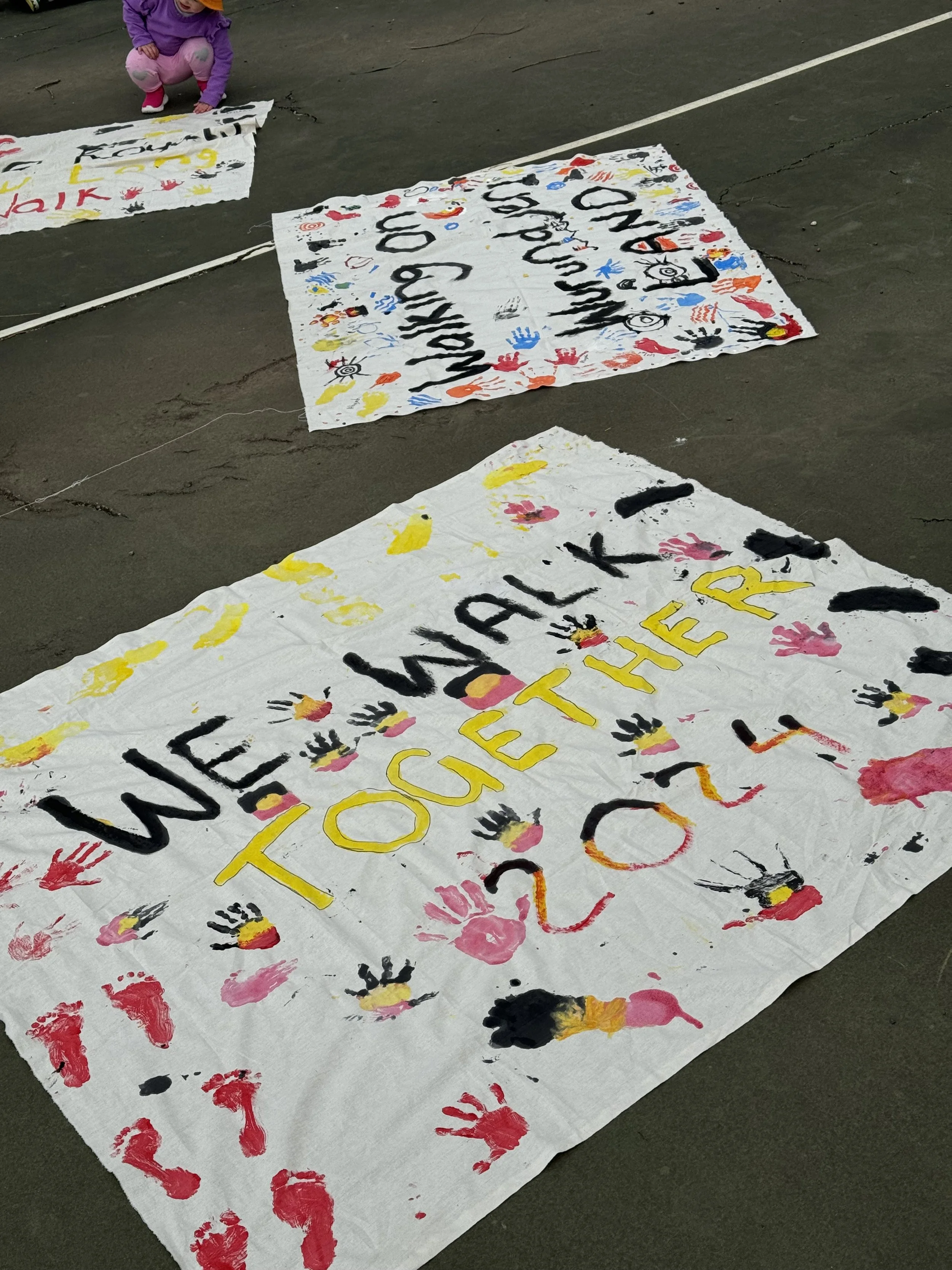 Children's artwork on large white sheets of paper with colorful handprints, painted words, and drawings on a concrete floor.