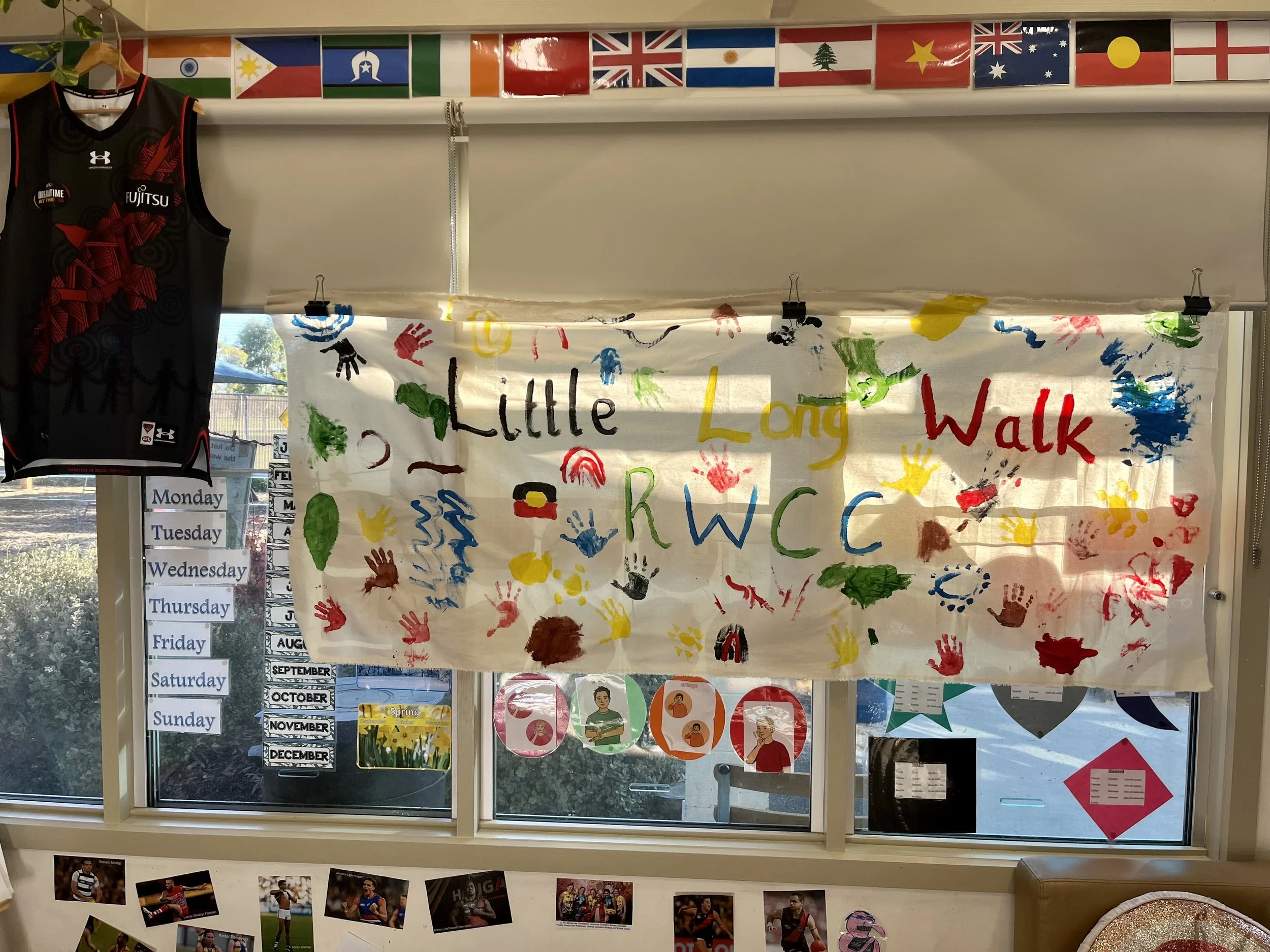 A classroom window decorated with a colorful banner with handprints and the words 'Little Long Walk'. A row of small international flags runs across the top of the window. To the left, there's a basketball jersey hanging and a weekly schedule poster 