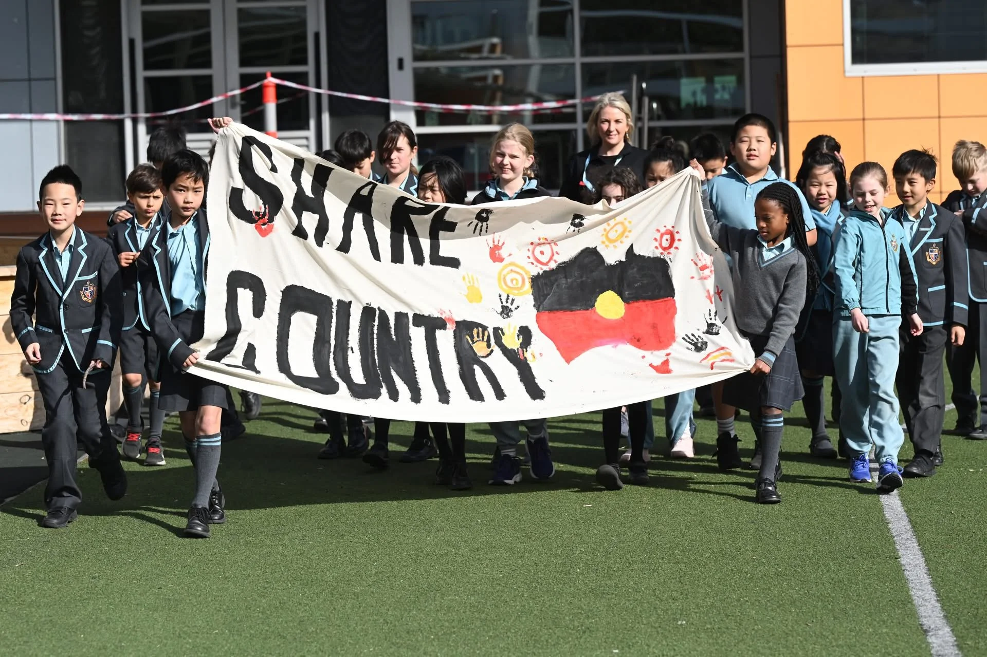 Group of children holding a banner that reads 'Save Our Country' with artwork of the Australian flag and red, yellow, and black colors, walking outdoors in front of a modern building.