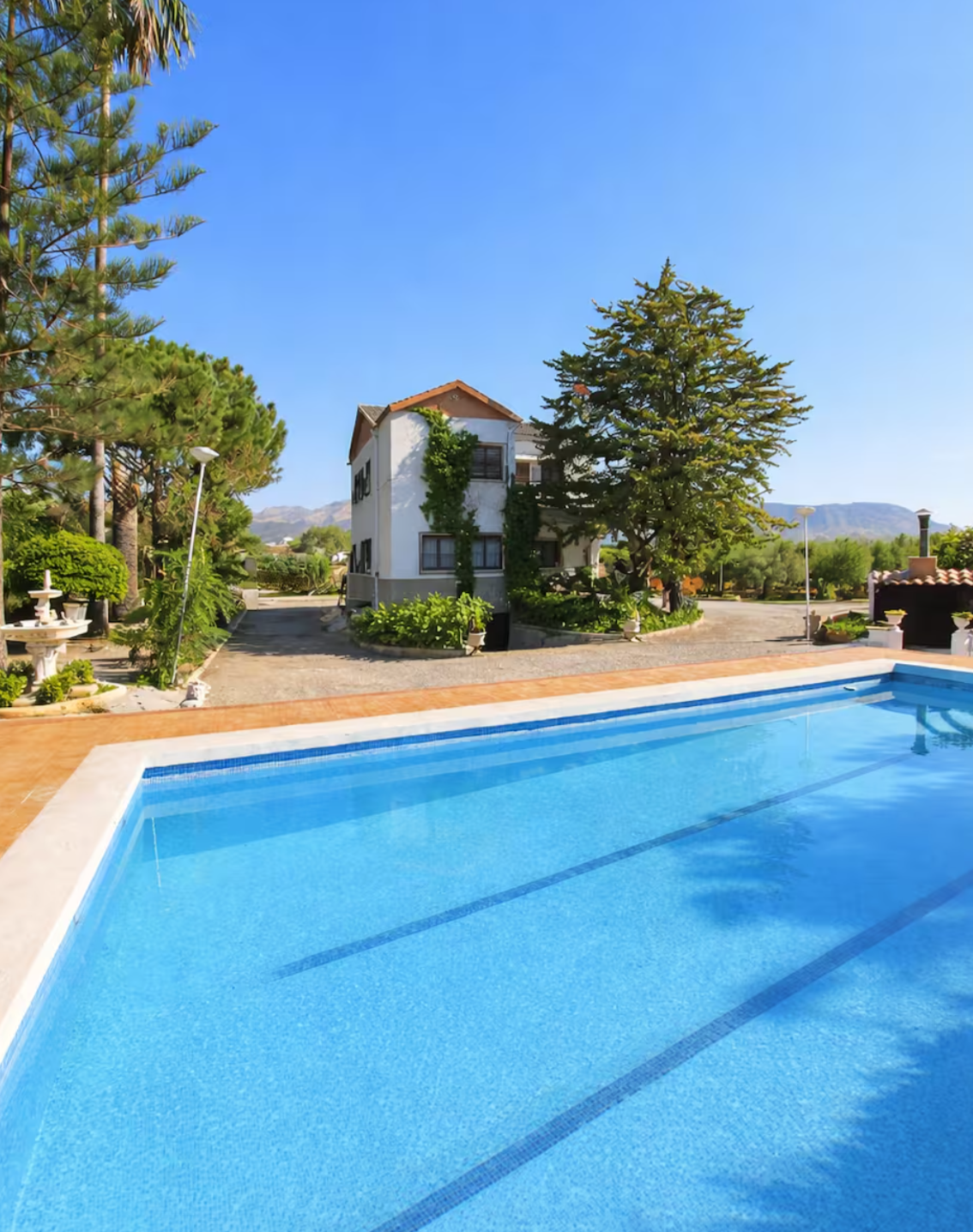 A backyard swimming pool with blue water in a sunny outdoor setting, with a white house, green trees, and a clear blue sky in the background.