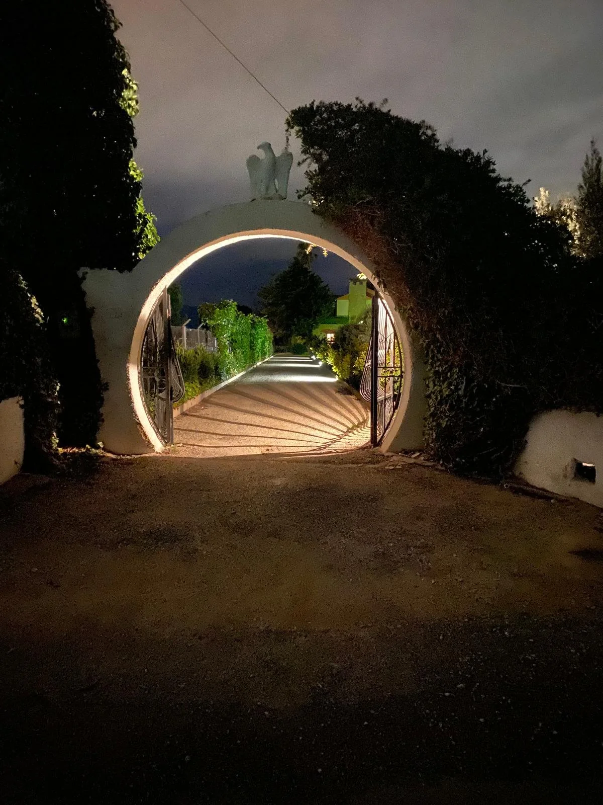 Night view of a circular gate with an eagle sculpture on top, illuminated from below, with a photo of a driveway, plants, and trees in the background.