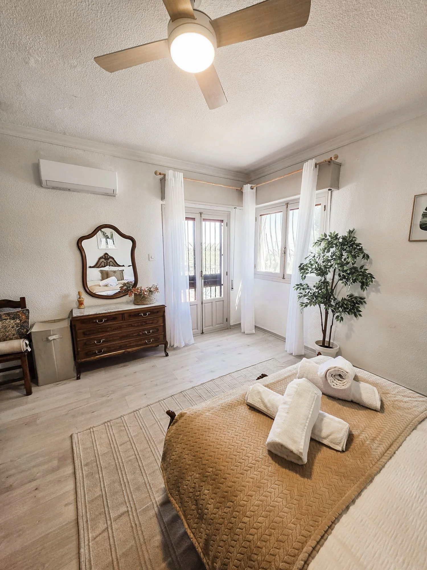 A bedroom with a bed covered with a textured beige blanket, folded white towels on top, a wooden dresser with a mirror, a potted plant, white curtains, and a window with a balcony door. There is a ceiling fan with lights and an air conditioner on the wall.