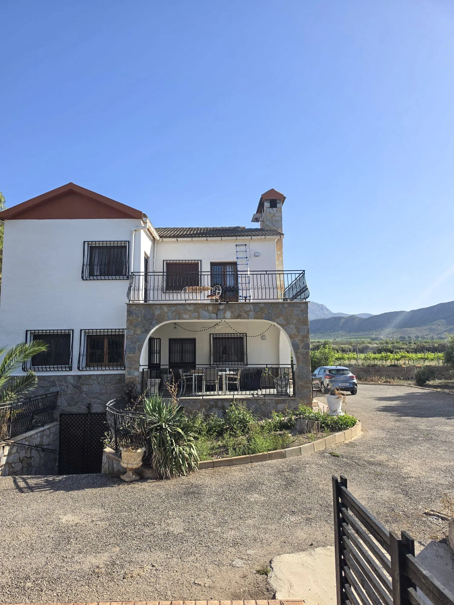 A two-story white house with a balcony, black railings, and a stone base, set against a backdrop of mountains and a clear blue sky, with a parking area and some plants in the foreground.