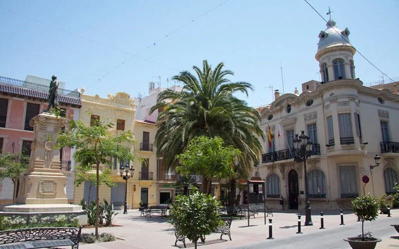 A city square with a statue, palm tree, small trees, benches, and historic buildings with European architectural style.