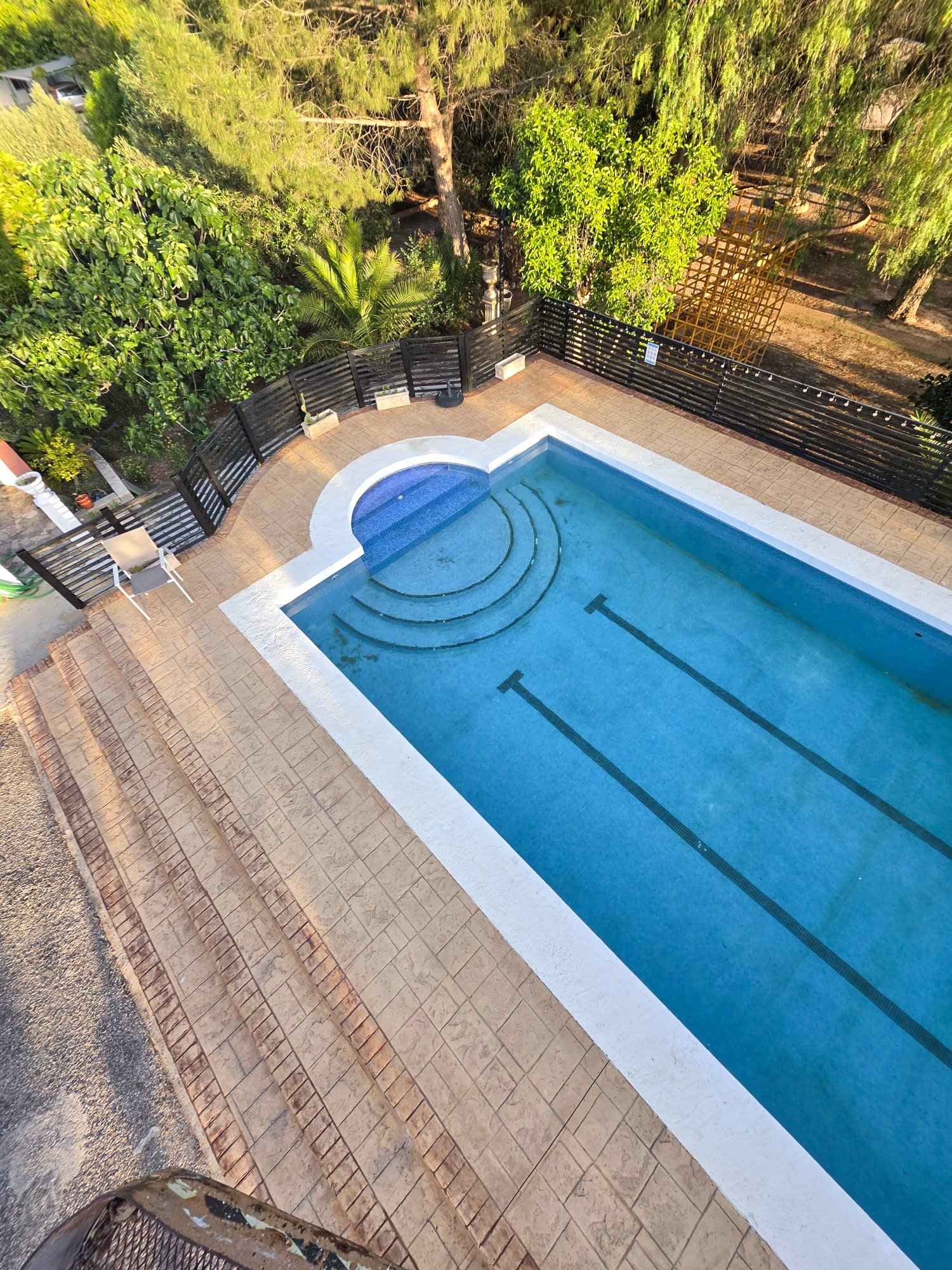An aerial view of a swimming pool with steps, surrounded by a tan tiled deck, black fence, and lush green trees.