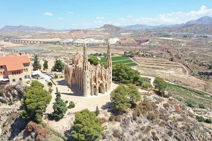 A large Gothic-style church with tall spires situated on a hill in a semi-arid landscape. There are trees and buildings nearby, with mountains and a valley in the background under a partly cloudy sky.
