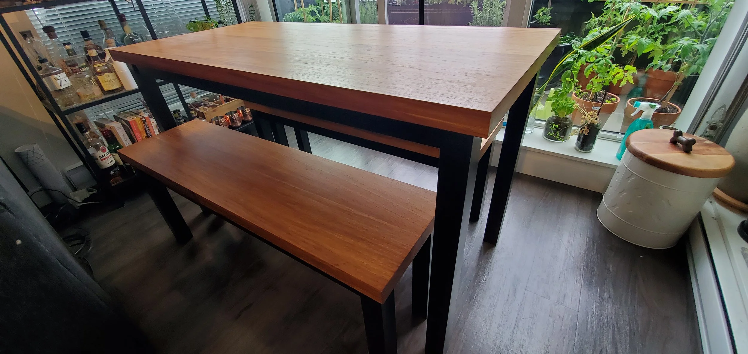 A wooden dining table with a matching bench in a room with a large window, potted plants on the windowsill, and a bookshelf with bottles and books.