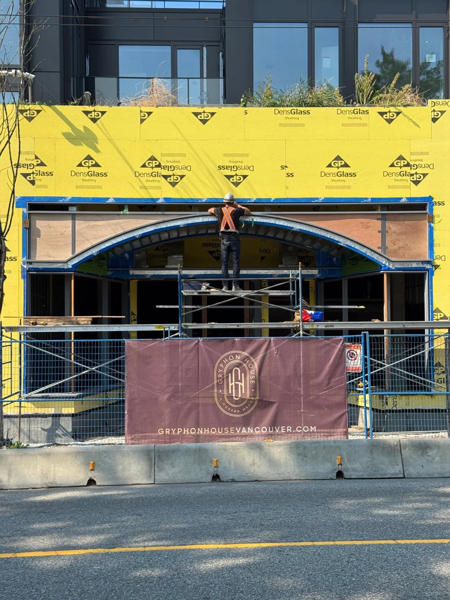 Construction worker standing on scaffolding installing an arched decorative window frame on a building under construction. The building exterior is covered with yellow insulation wrap labeled 'DensGlass.' There is a banner for Gryphon House Vancouver