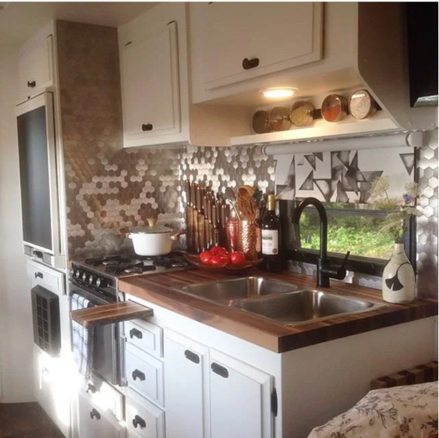 Kitchen with white cabinets, wooden countertop, stainless steel sink, mosaic tile backsplash, and a window with greenery outside. Various kitchen utensils, a bowl, tomatoes, wine bottle, and a decorative vase are on the counter.