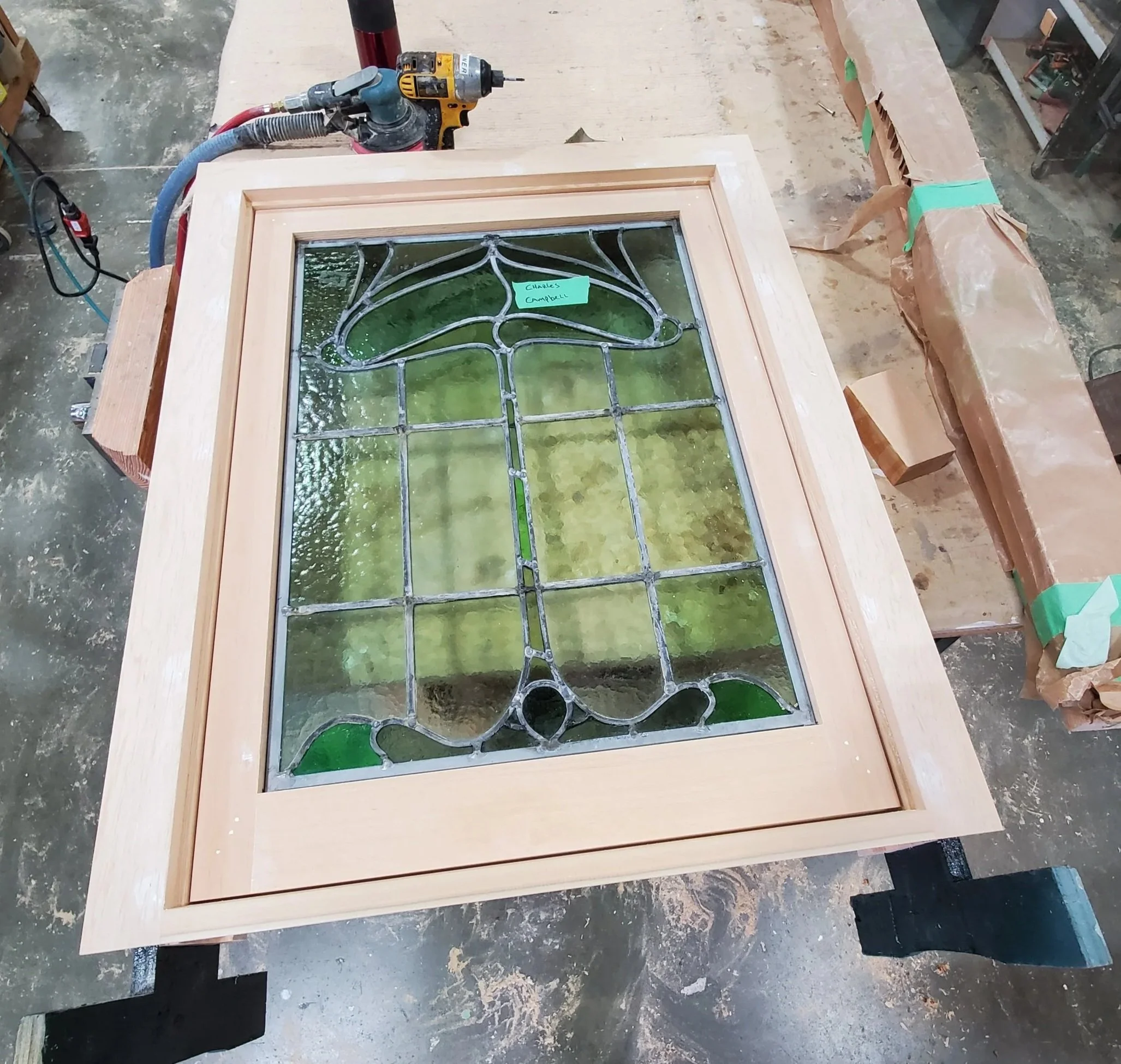 Stained glass window with green and clear glass pieces and intricate lead framework, resting on a wooden frame in a workshop.