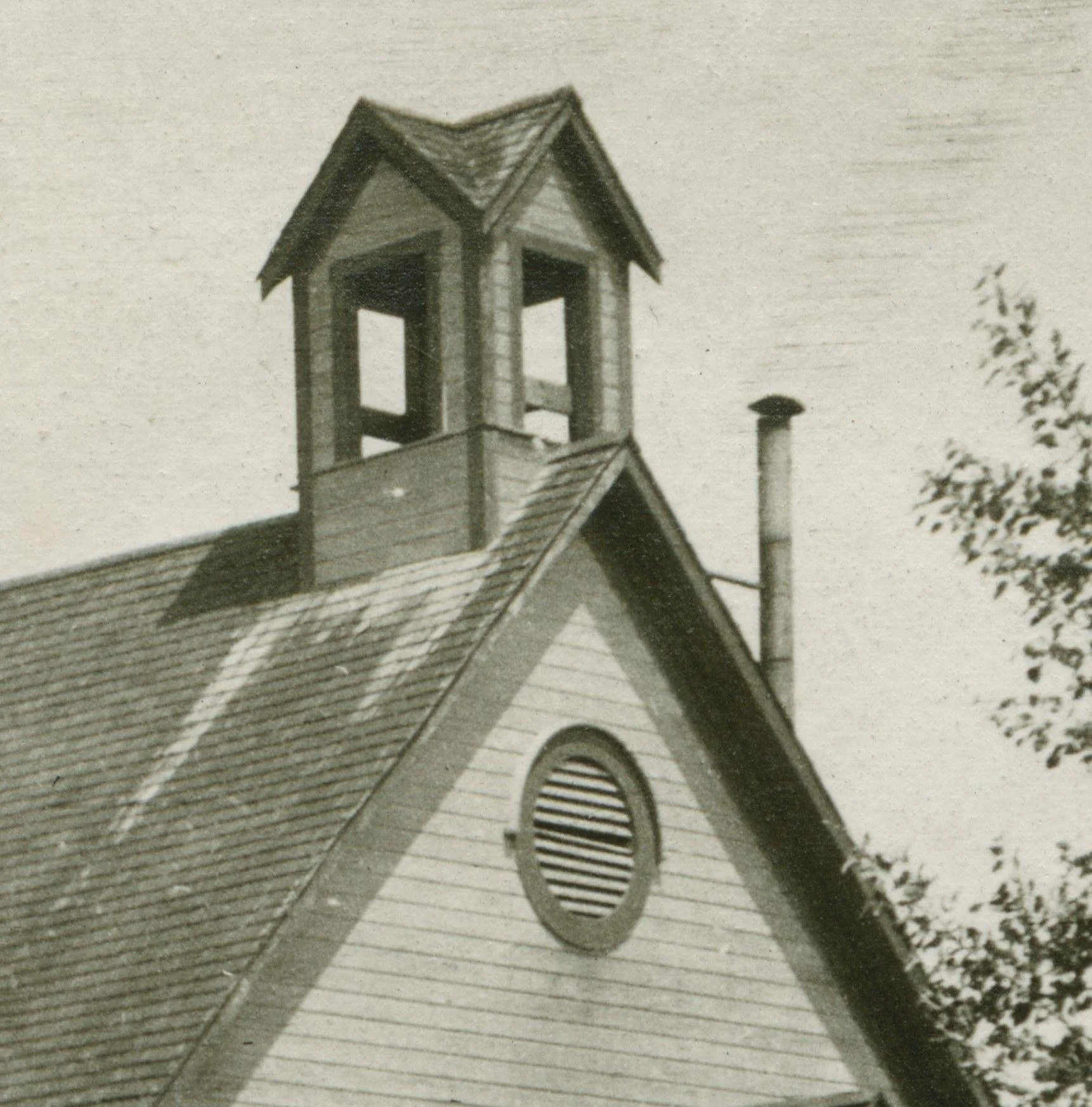 Black and white photo of the roof of a church or similar building, showing a small bell tower with open windows on each side and a shingled roof. A chimneystack is visible near the steeple, and part of a tree is on the right.