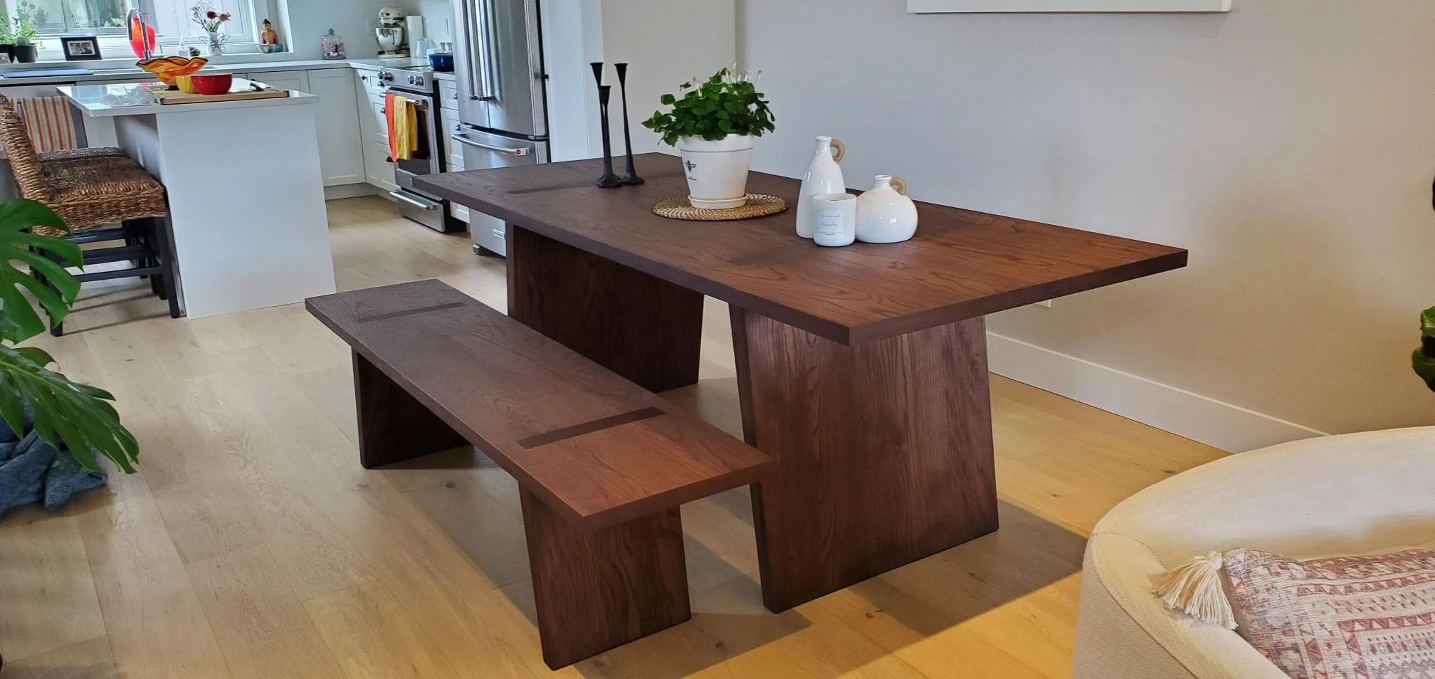 A wooden dining table with a matching bench, decorated with potted plant and white vases, in a modern kitchen and living room setting.