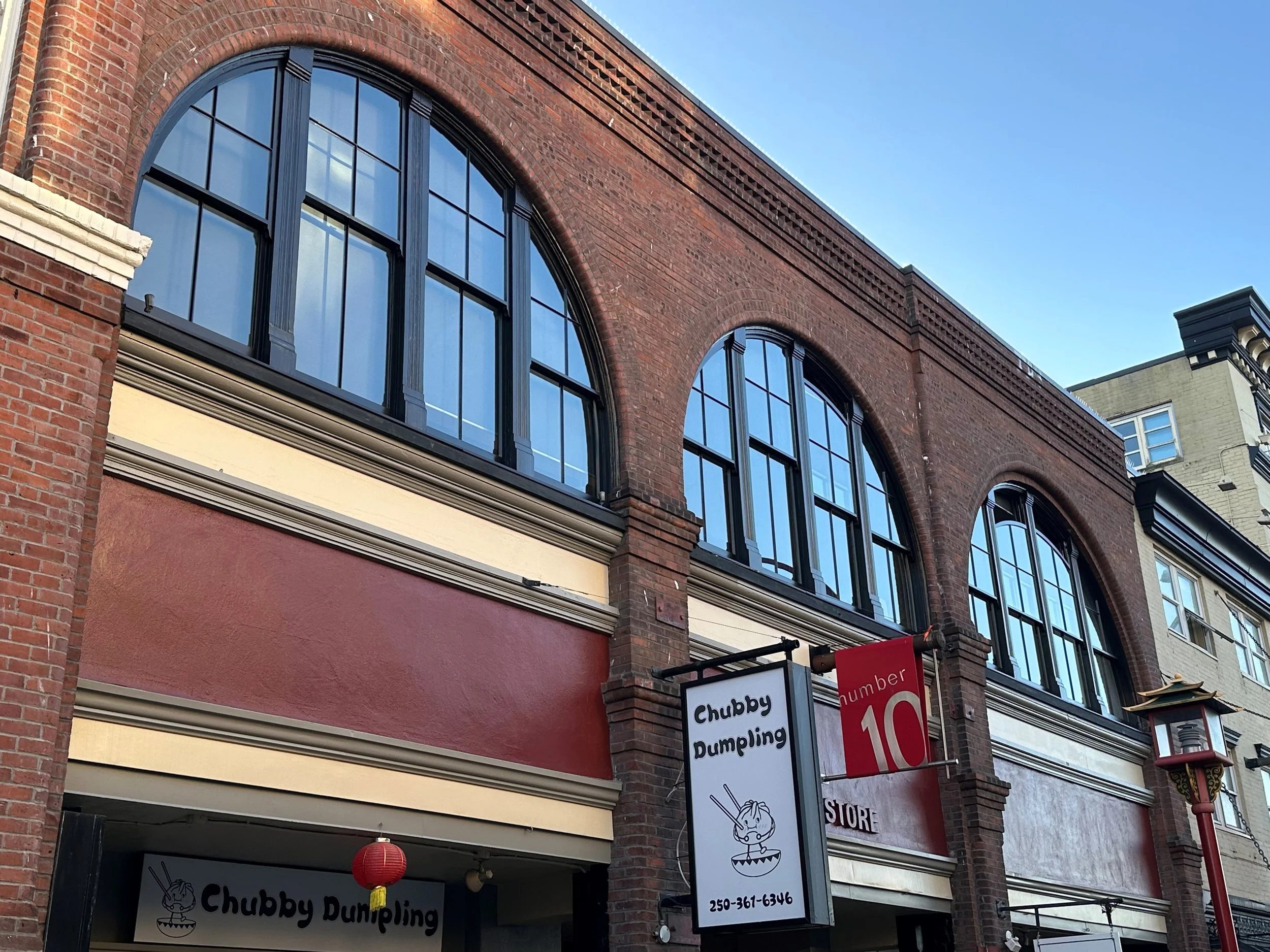 A storefront named Chubby Dumpling with a sign showing a cartoon dumpling character eating with chopsticks and a phone number, located on a brick building with large arched windows, a red and beige awning, and a decorative street lamp, with another b