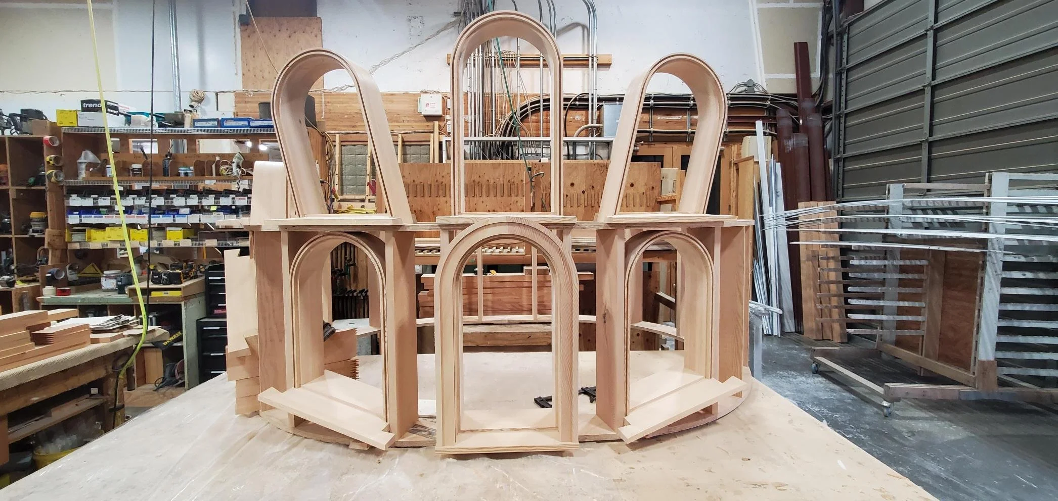 Wooden architectural model or sculpture in a woodworking shop, featuring arches and curved structures, set on a workbench with various woodworking tools and materials in the background.