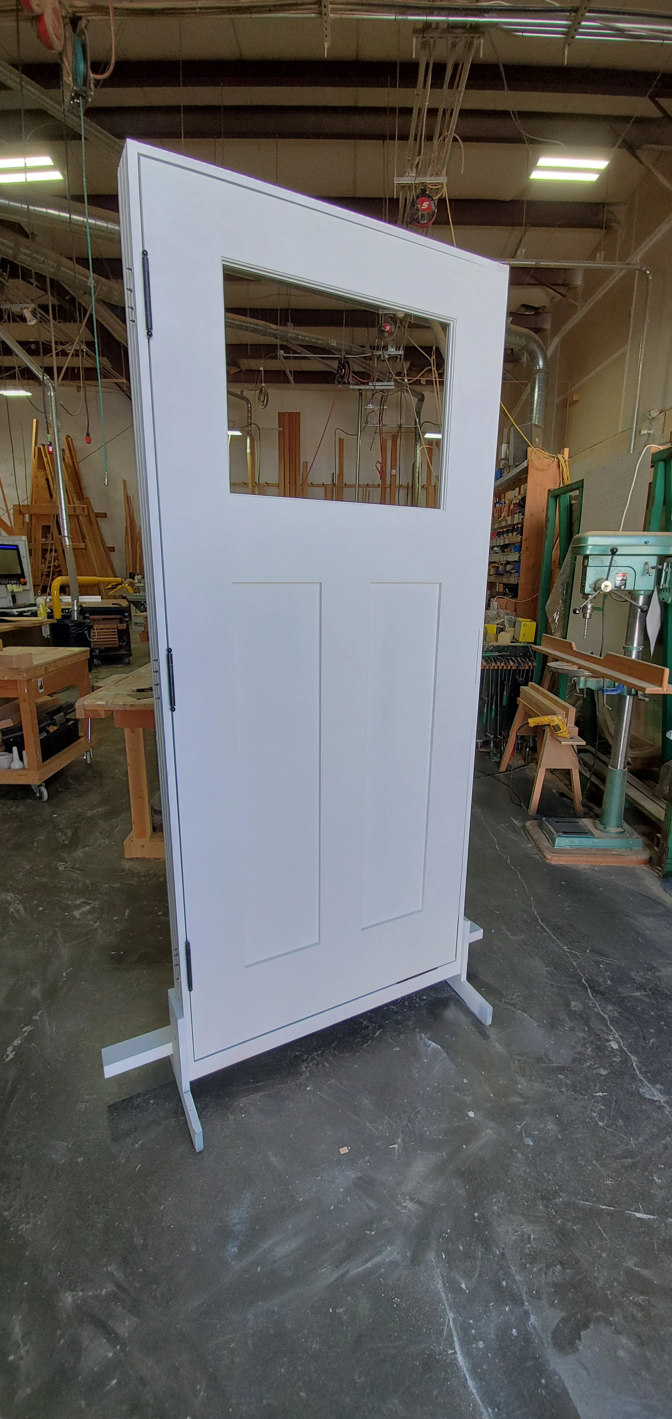 White interior door with a window, standing on a workshop floor surrounded by woodworking tools and equipment.