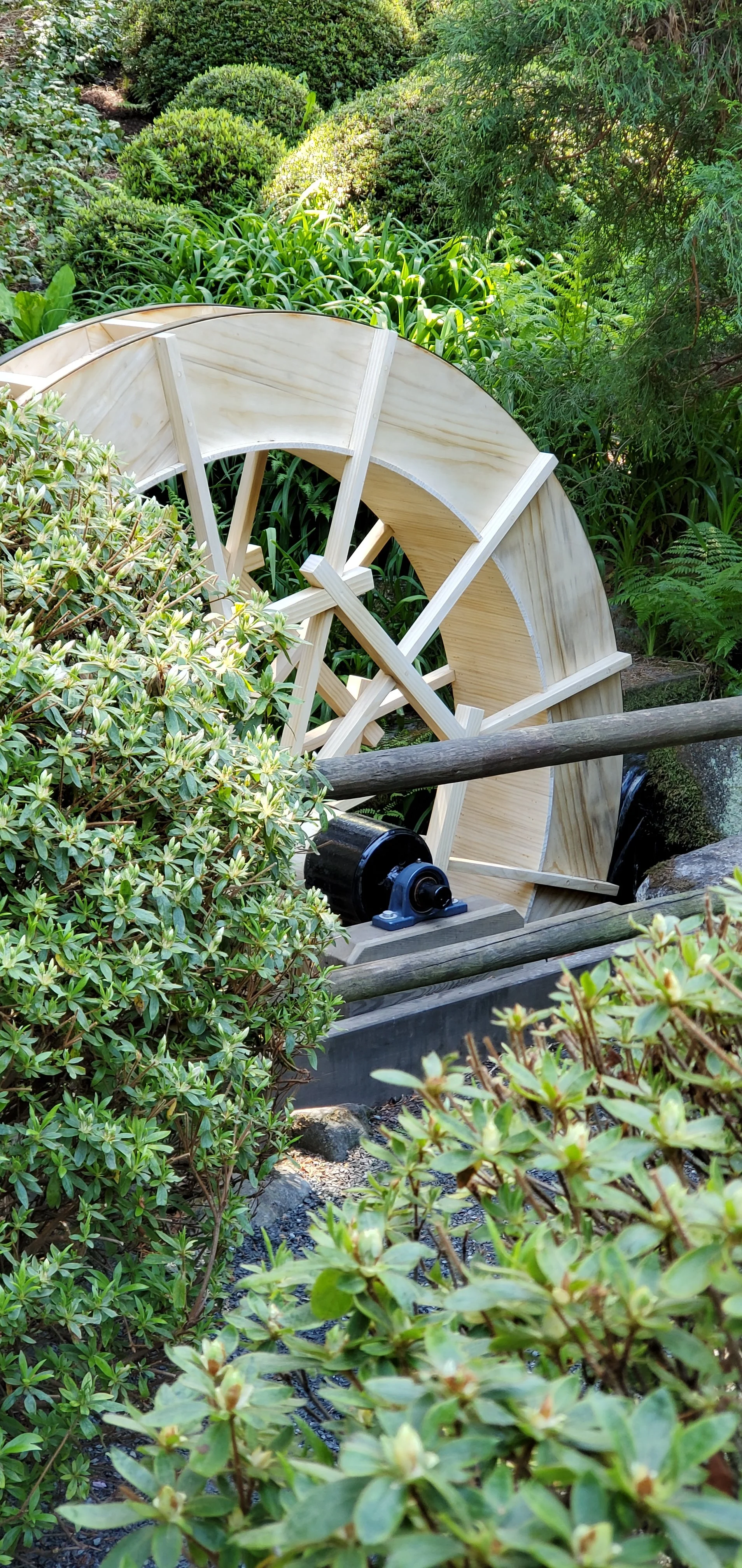 A wooden waterwheel in a lush green garden surrounded by various bushes and plants.