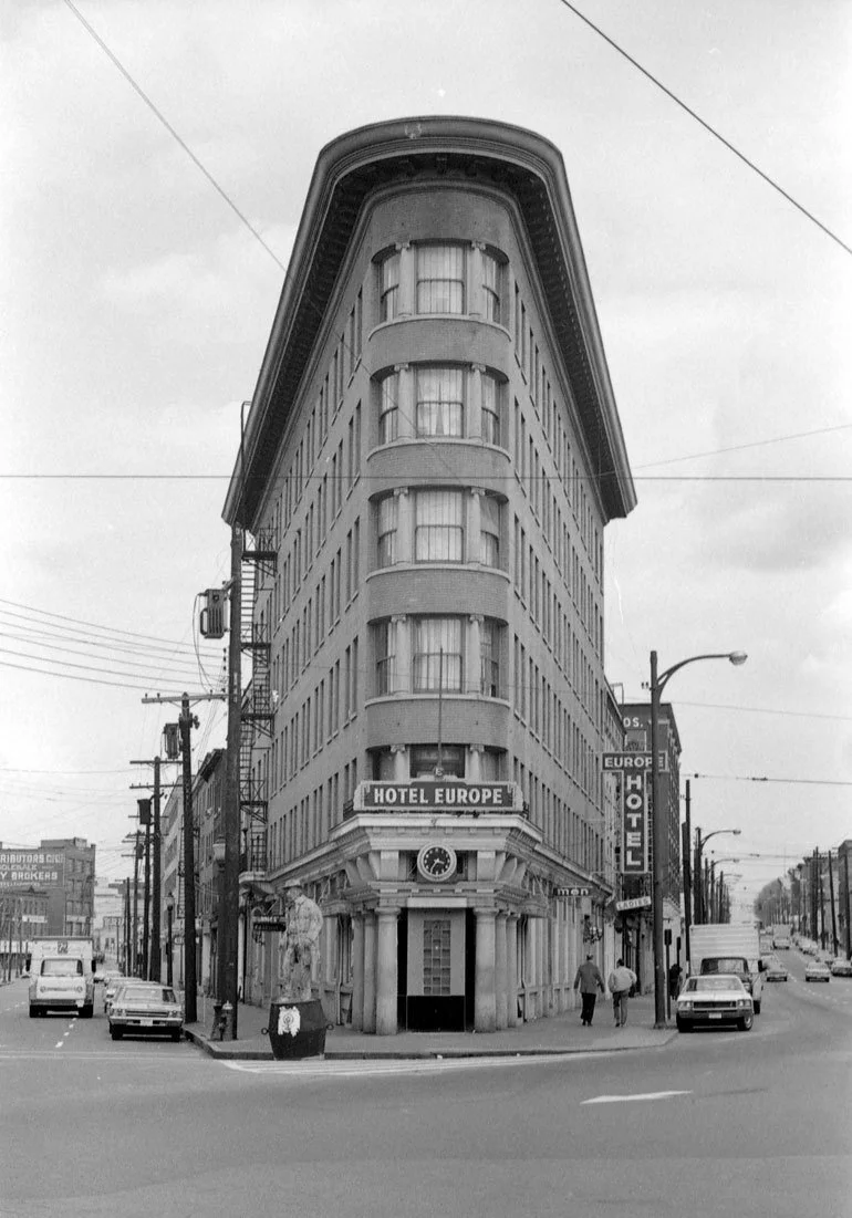 A black and white photo of a tall, narrow, seven-story building on an intersection, labeled as Hotel Europe, with a rounded corner and large windows. Cars are parked on the street, and pedestrians are walking nearby.