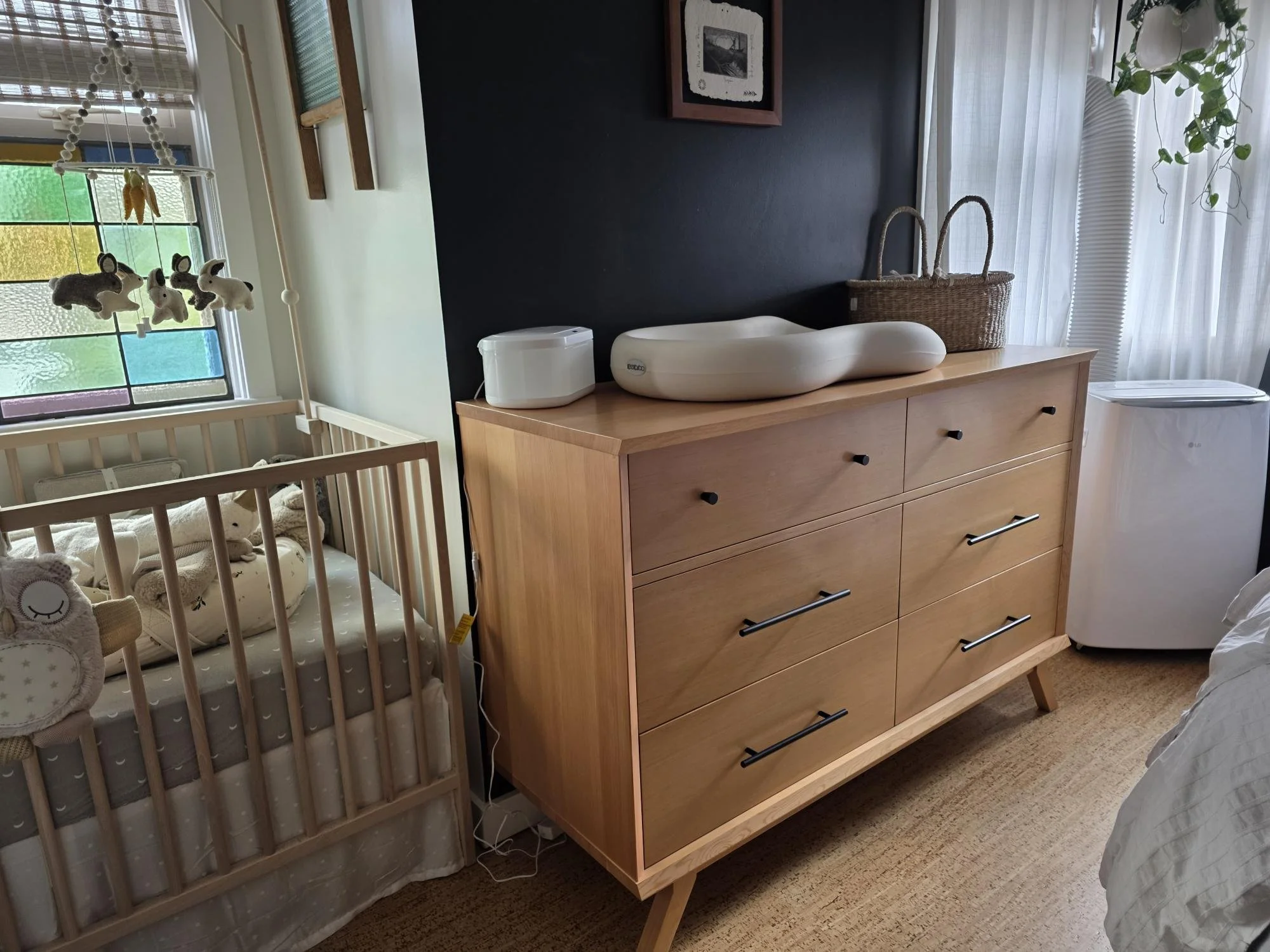 A wooden dresser with six drawers, a changing pad on top, a rice steamer, and a woven basket inside a room with a black accent wall, sheer curtains, and a crib with plush toys and stuffed animals.