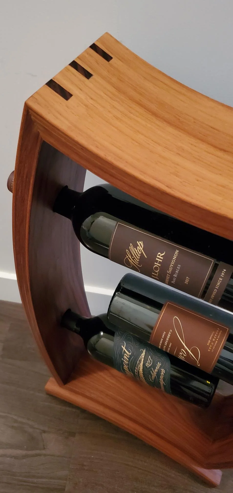 A wooden wine rack holding three bottles of red wine, positioned against a neutral-colored wall and placed on a wooden floor.