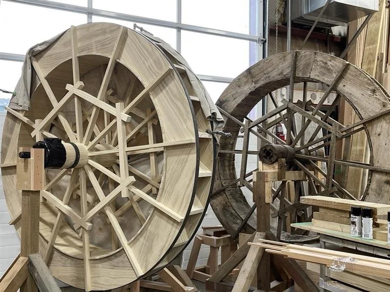 Two large wooden wheels under construction in a woodworking shop, with various tools and materials around.
