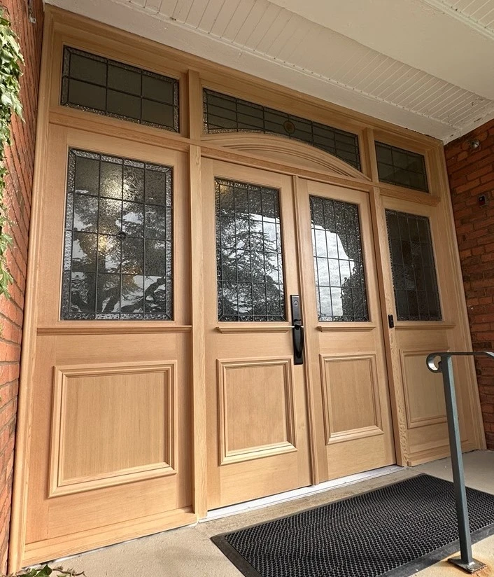 New wooden front door with decorative glass panels, black handles, and a black doormat.