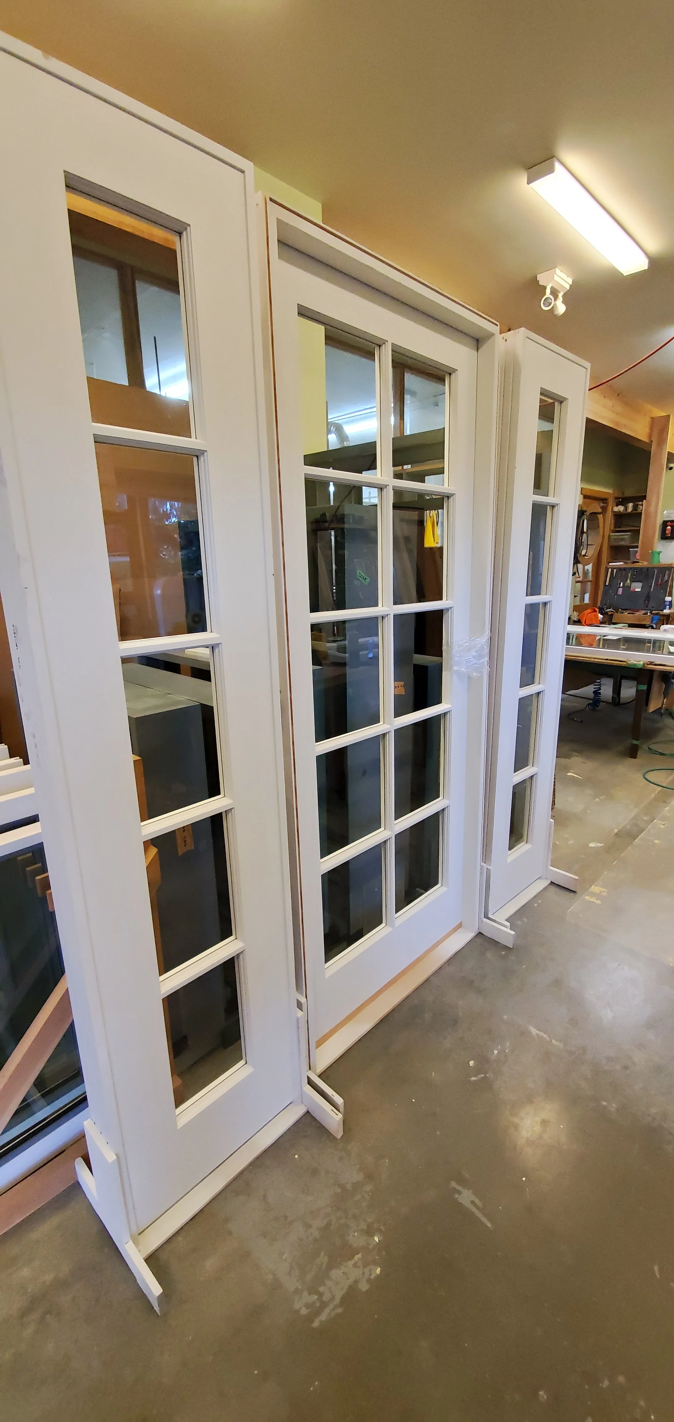 Three white framed glass-paneled doors in a woodworking workshop, standing upright on a concrete floor.