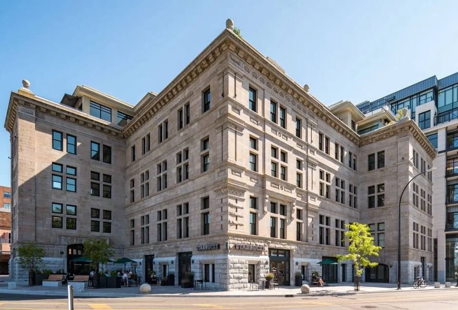 Street view of a large, historic-style building with Starbucks and other storefronts at street level, featuring multiple stories, numerous rectangular windows, and decorative architectural details.