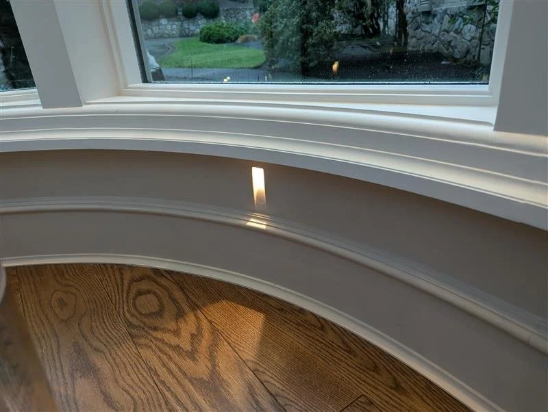 View of a bay window with white trim, showing a reflection of a light on the window frame and a glimpse of a yard with bushes and a stone stair outside.