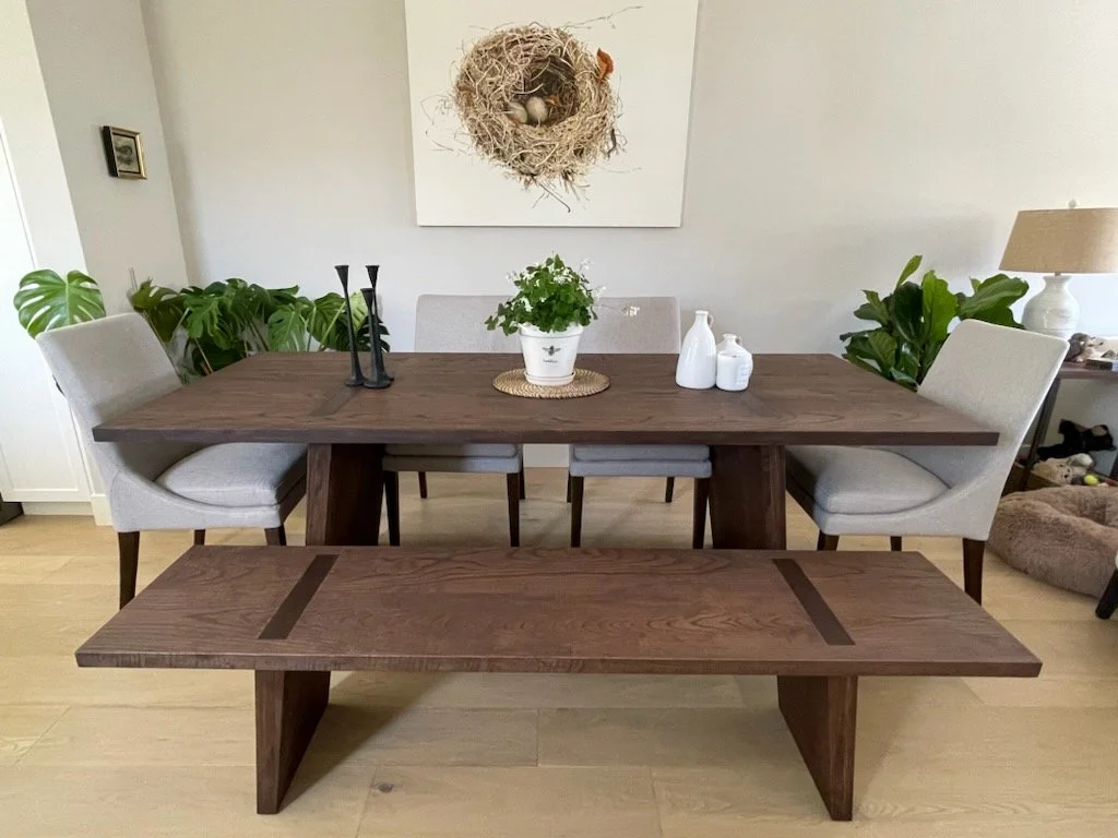 A dining room with a wooden table, six beige upholstered chairs, a matching wooden bench, green plants, a white piece of art with a nest and bird in it on the wall, a white potted plant, three white decorative bottles, a beige table lamp, and a small