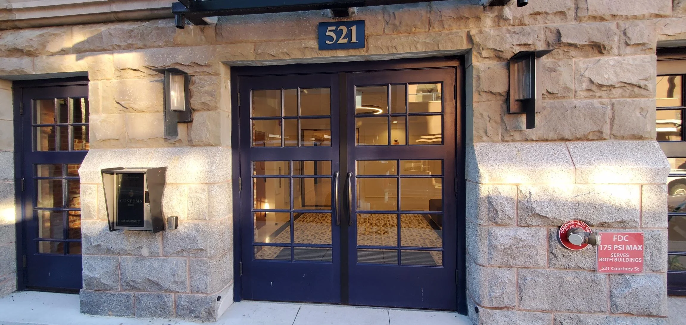 The entrance to a building with double glass doors and a black metal frame. A blue number 521 is above the doors. There are two wall-mounted lights on either side of the door, and a red fire alarm with a label that states 175 PSI max for both buildin