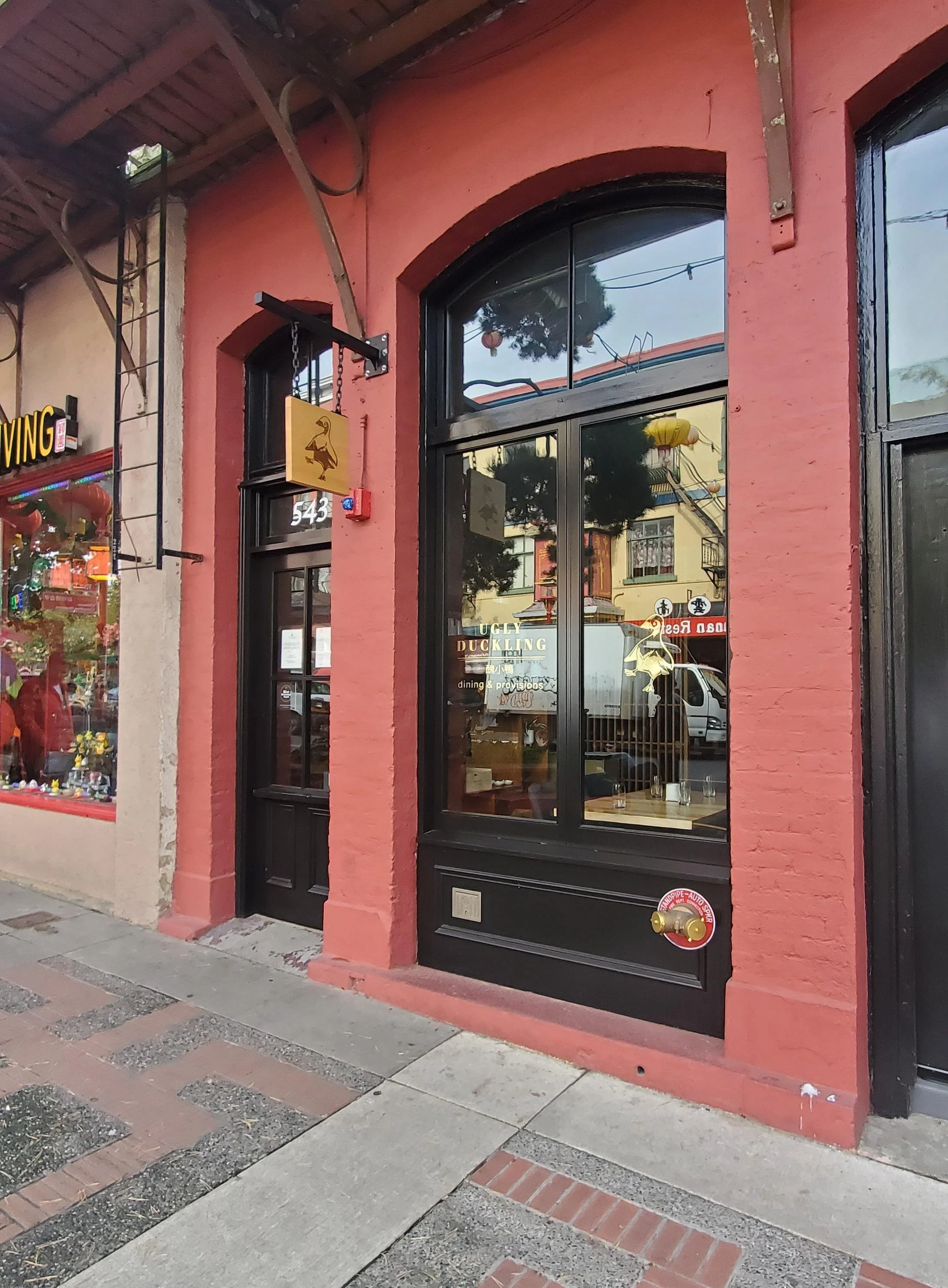 The exterior of a restaurant with a pink wall, black door, large window, hanging sign with a duck silhouette, and brick sidewalk.