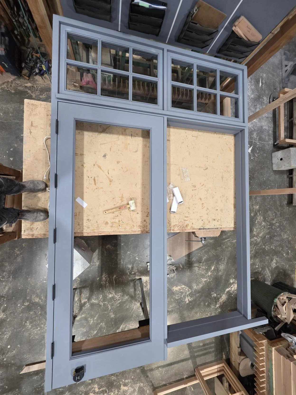 Blue unfinished window frame with sidelights and grid windows on top, placed on a workbench in a workshop.