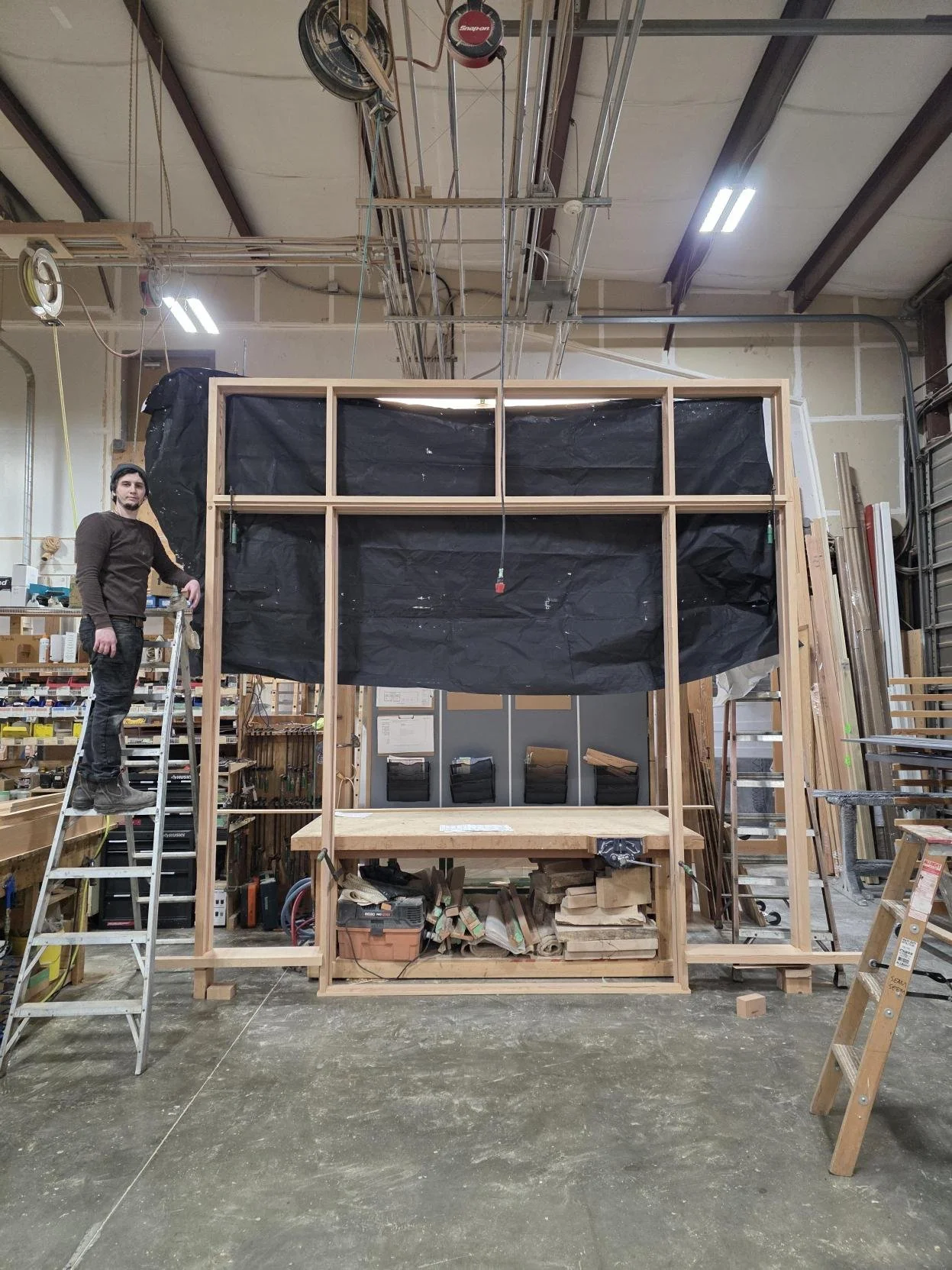 Person standing on a ladder next to a large wooden frame structure in a woodworking shop.