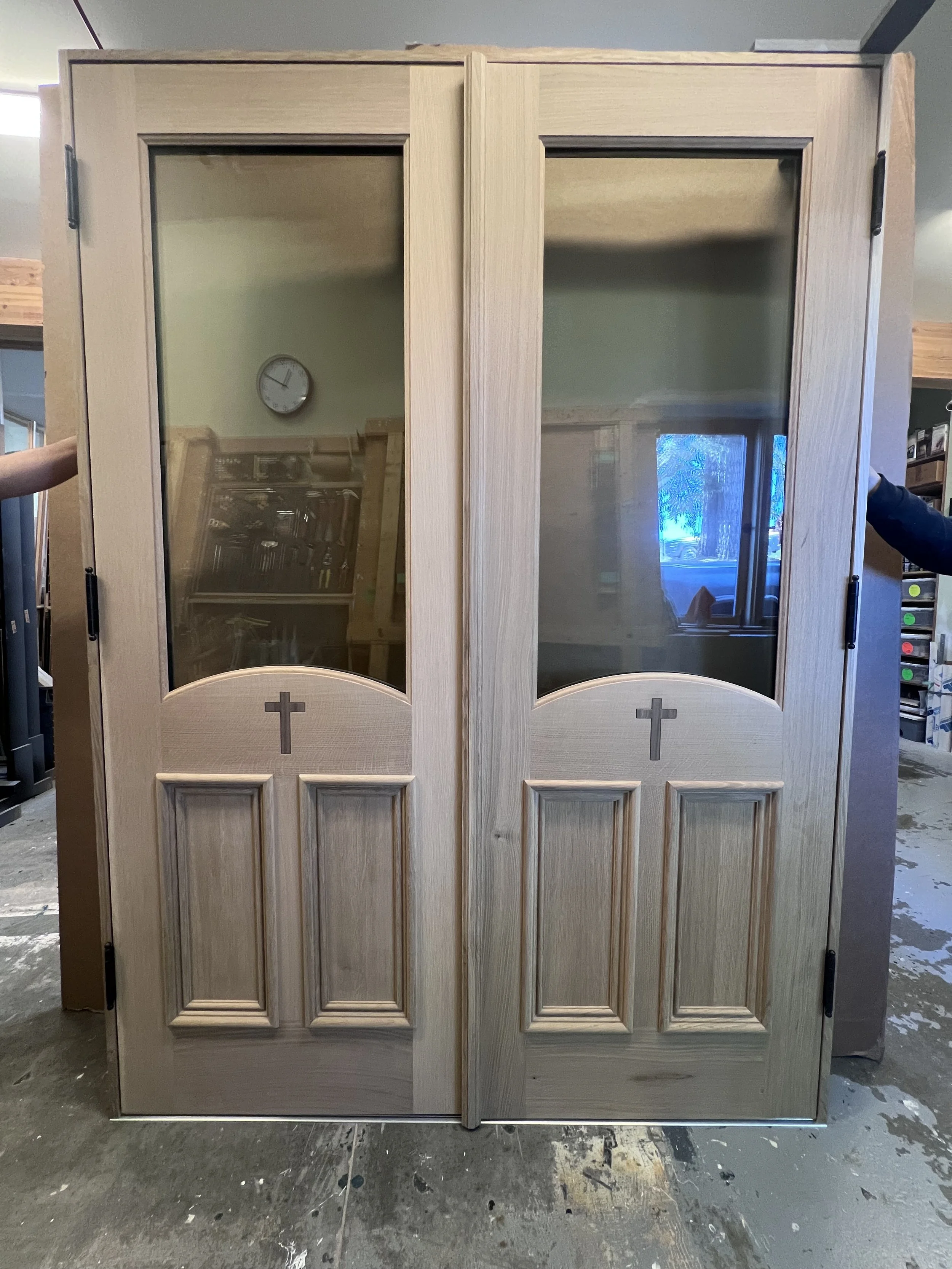 A pair of unfinished wooden church doors with glass panels and crosses carved in the lower part of each door.