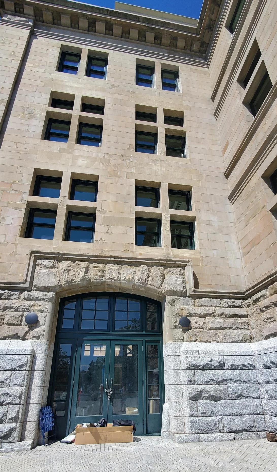 The image shows the facade of a historic stone building with arched and rectangular windows and an entryway with glass doors. Supplies are stacked outside the entrance.